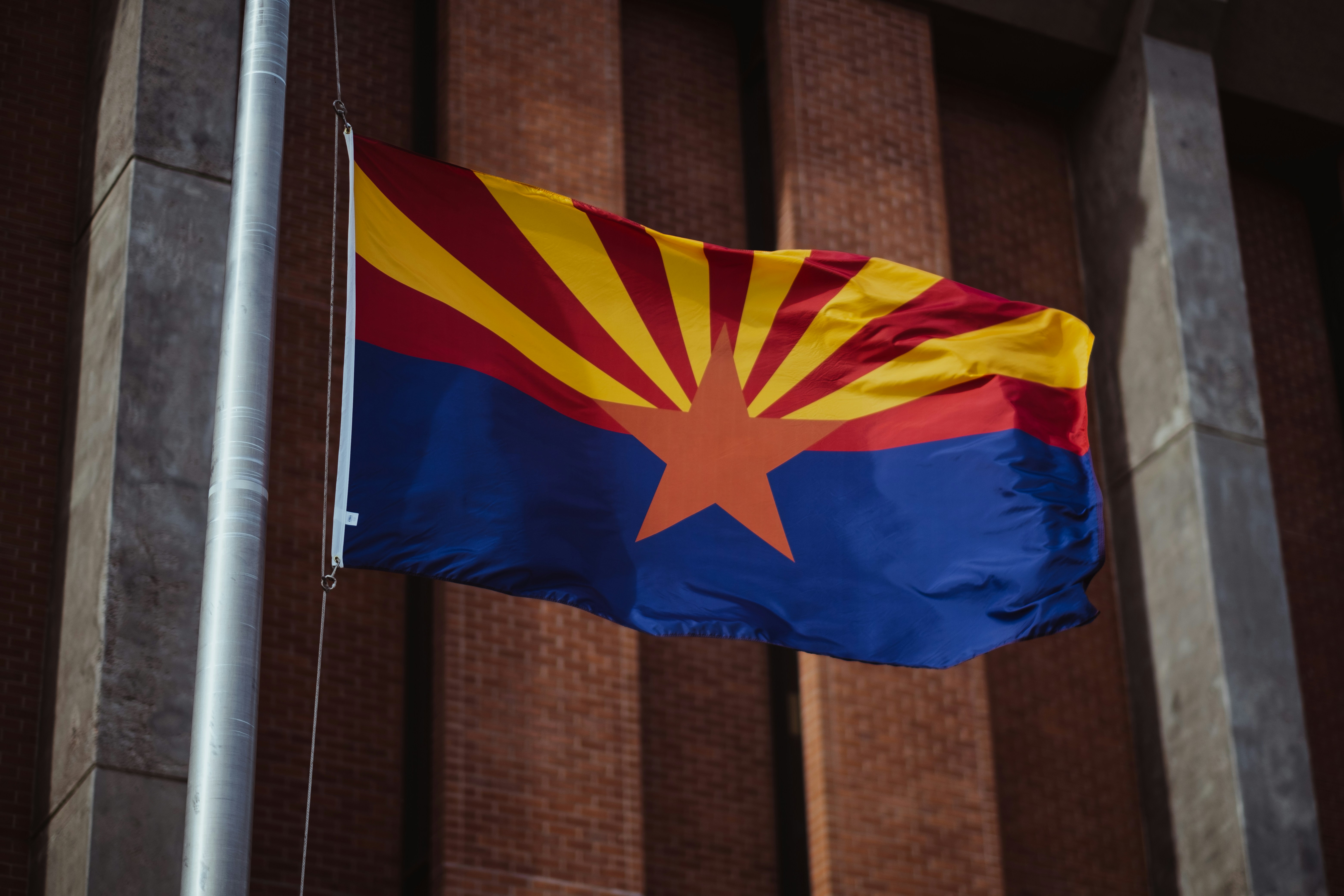 Arizona state flag with a copper star, red and yellow rays, and blue lower half flying on a pole in front of a brick building.