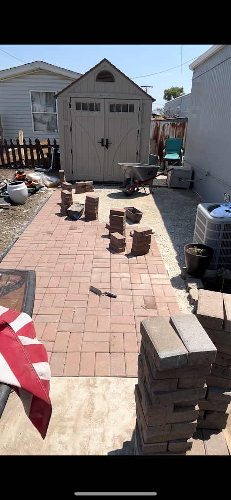 Backyard with partially installed red brick pathway leading to a beige storage shed, with stacks of bricks and a wheelbarrow nearby.