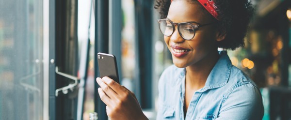 Young woman wearing glasses and a red headband smiling while looking at her smartphone near a window.