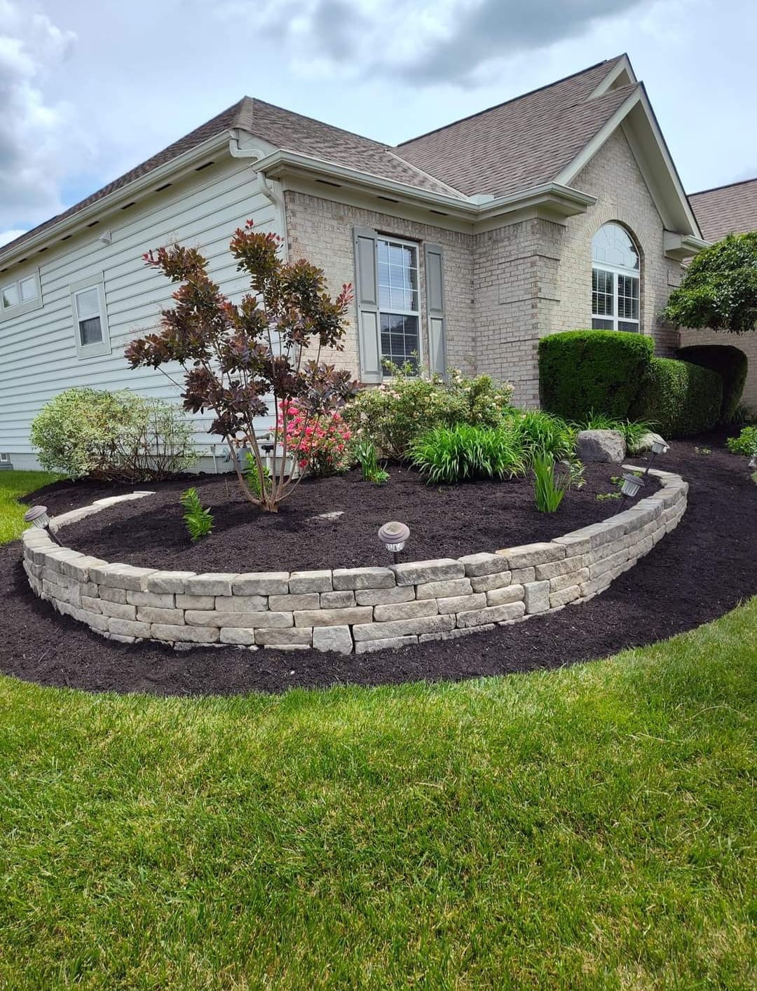 Curved stone retaining wall with fresh black mulch and various shrubs and plants in front of a brick and siding house.