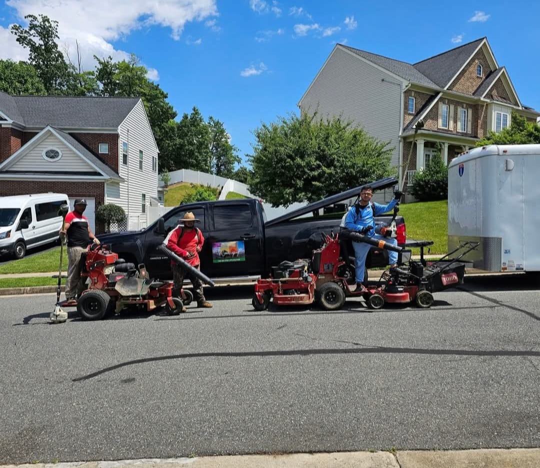 Three landscapers standing on a street with lawn care equipment, a black pickup truck, and a white trailer in a suburban neighborhood.