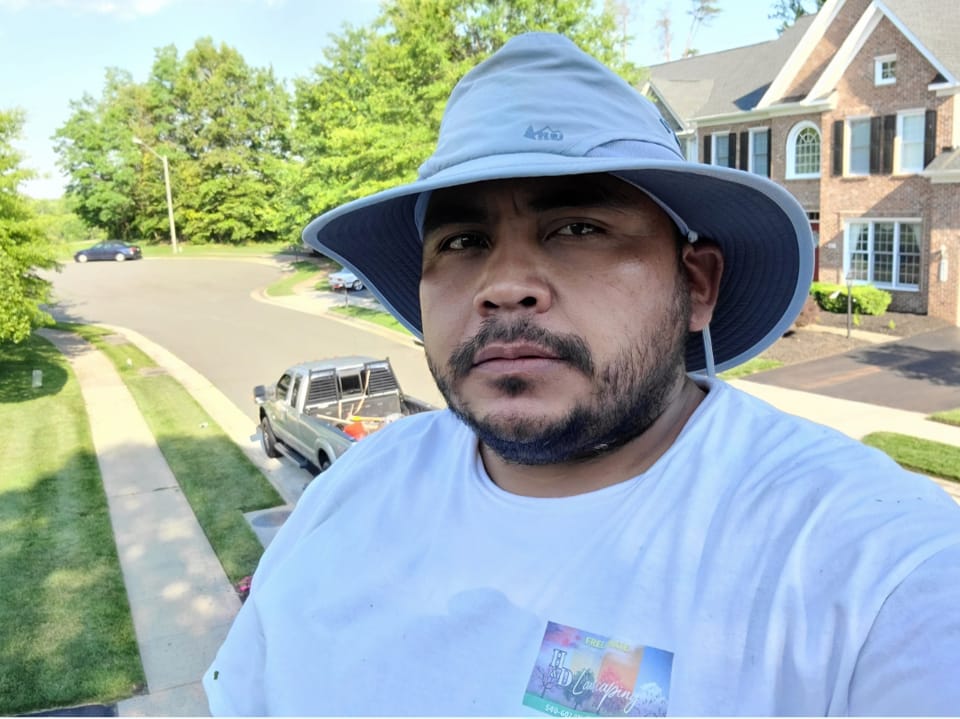 Man wearing a gray wide-brim hat and a white T-shirt standing outdoors in a suburban neighborhood with houses and green trees.