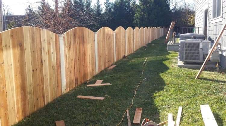 Newly installed wooden fence with curved tops running alongside a house with scattered wood pieces and tools on the grass.