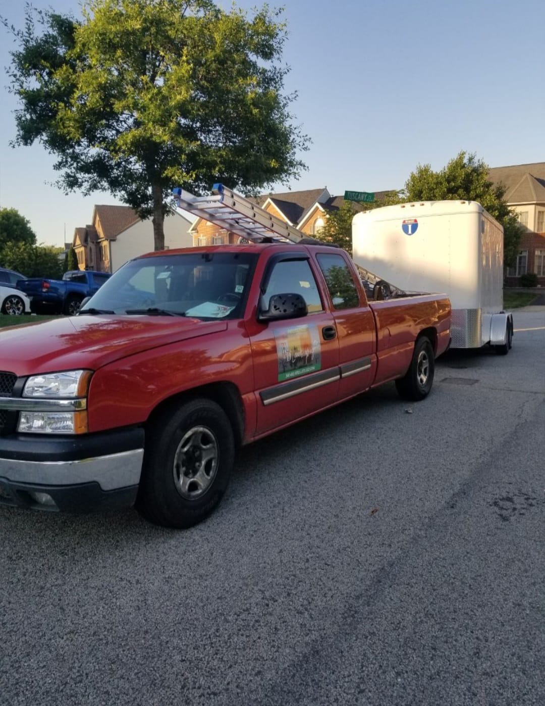Red pickup truck with ladders on the roof, towing a white enclosed trailer parked on a residential street.