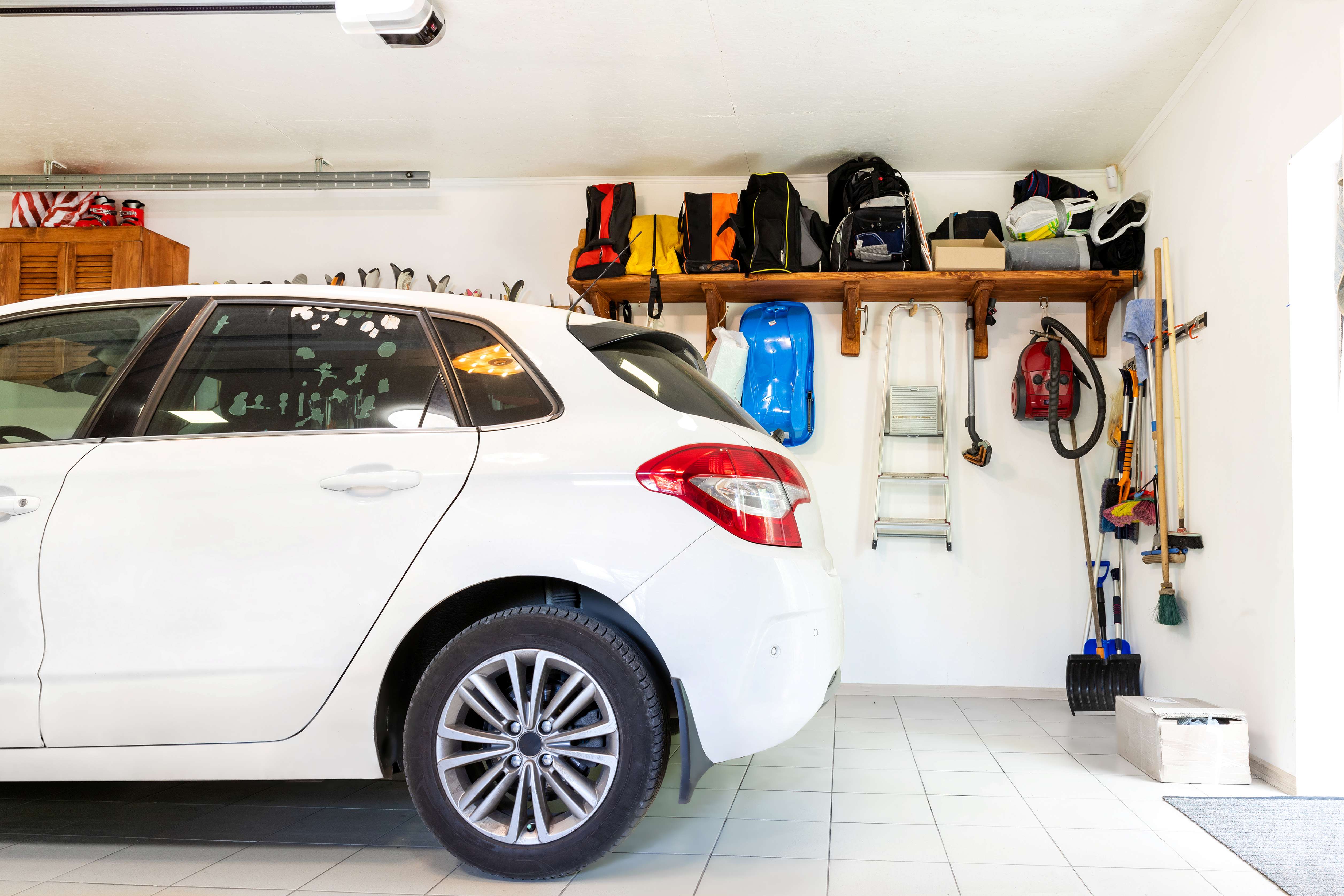 White car parked in a clean, organized garage with shelves holding bags and tools, and cleaning equipment hanging on the wall.