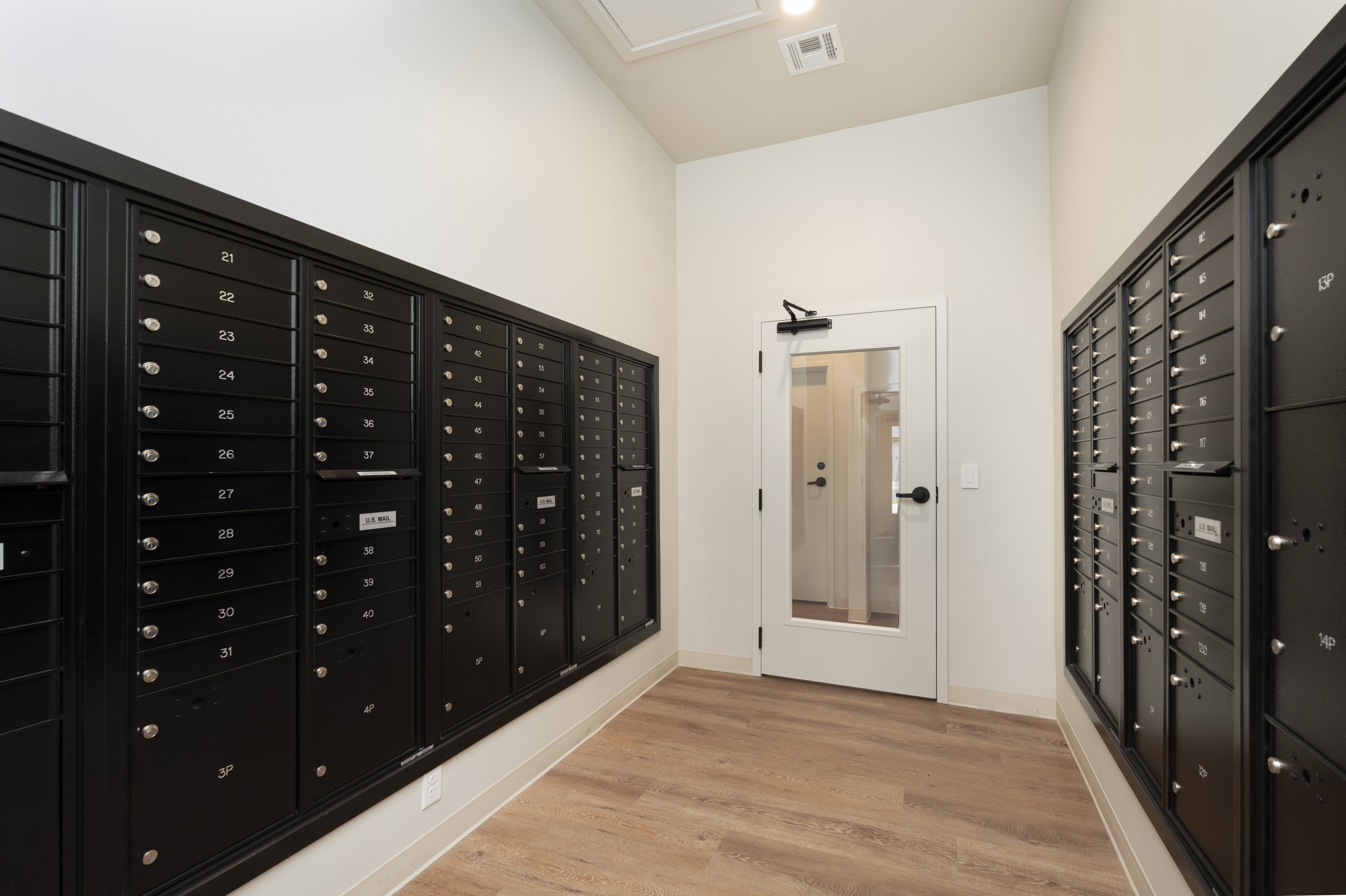 Interior mailroom with black numbered mailboxes on both walls and a white door with a window at the end.