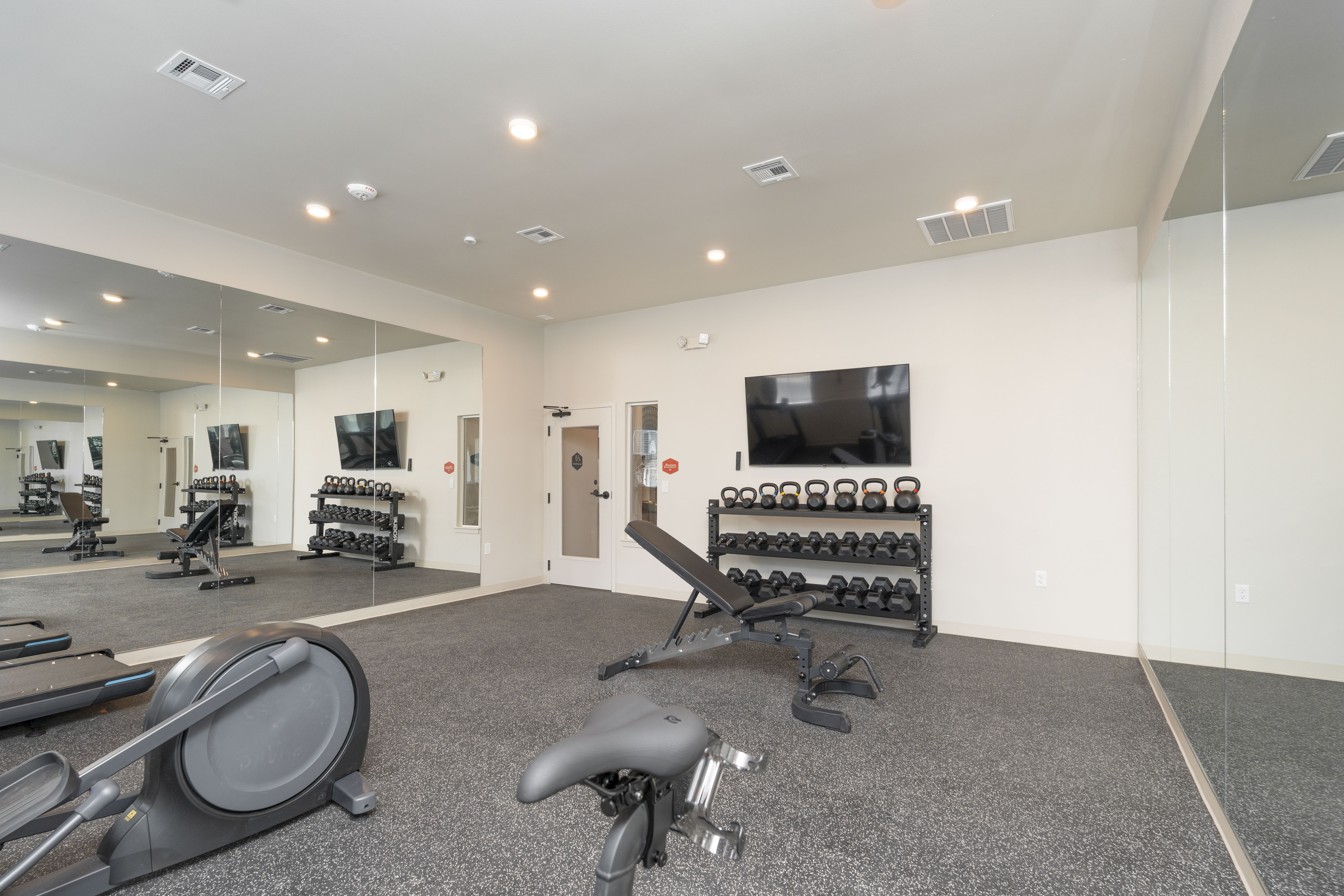 Modern gym room with adjustable bench, dumbbells, kettlebells, exercise bike, and mirrored walls.