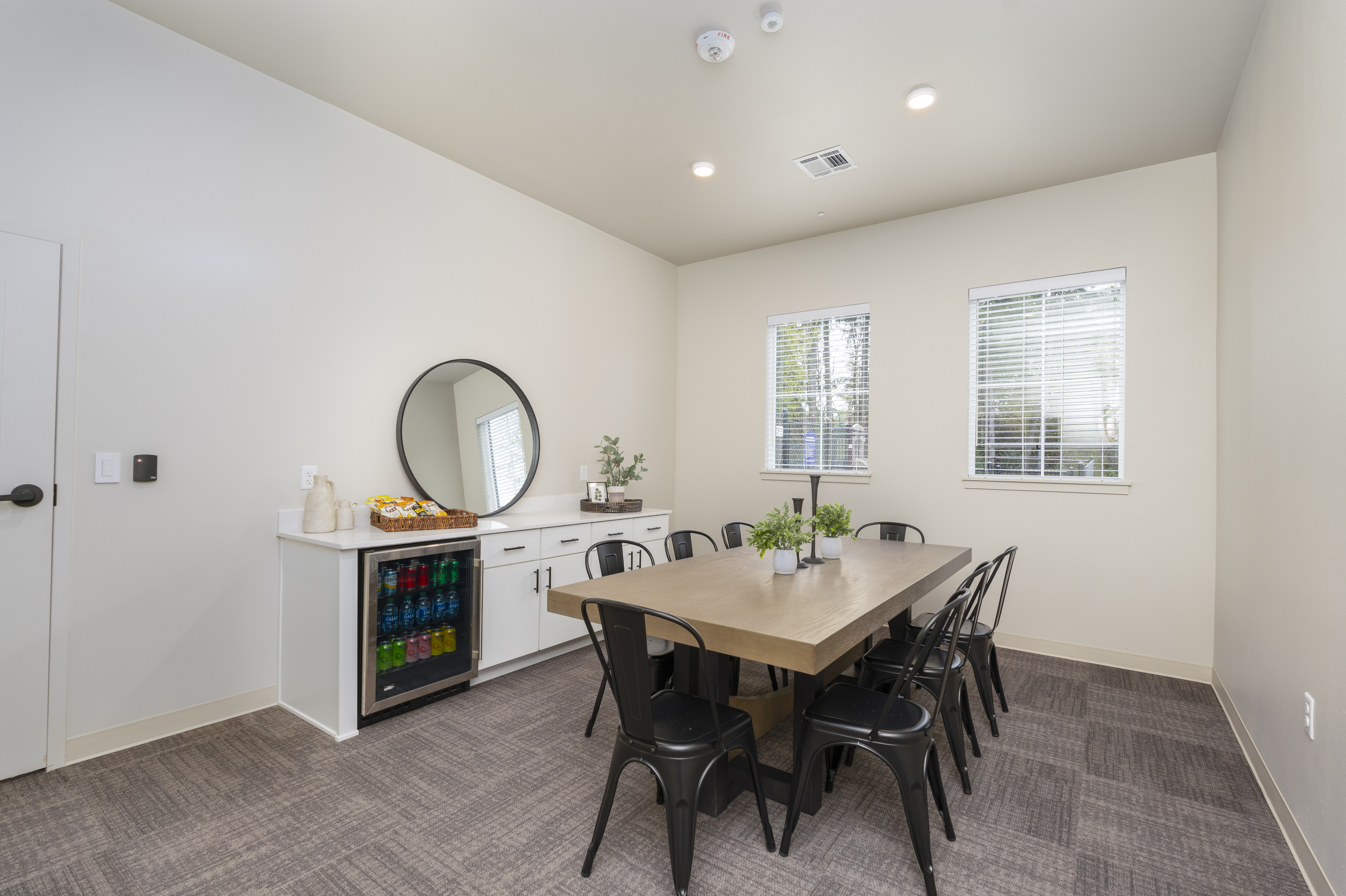Modern meeting room with a wooden table surrounded by black metal chairs, a cabinet with a mini fridge stocked with beverages, and two windows with blinds.