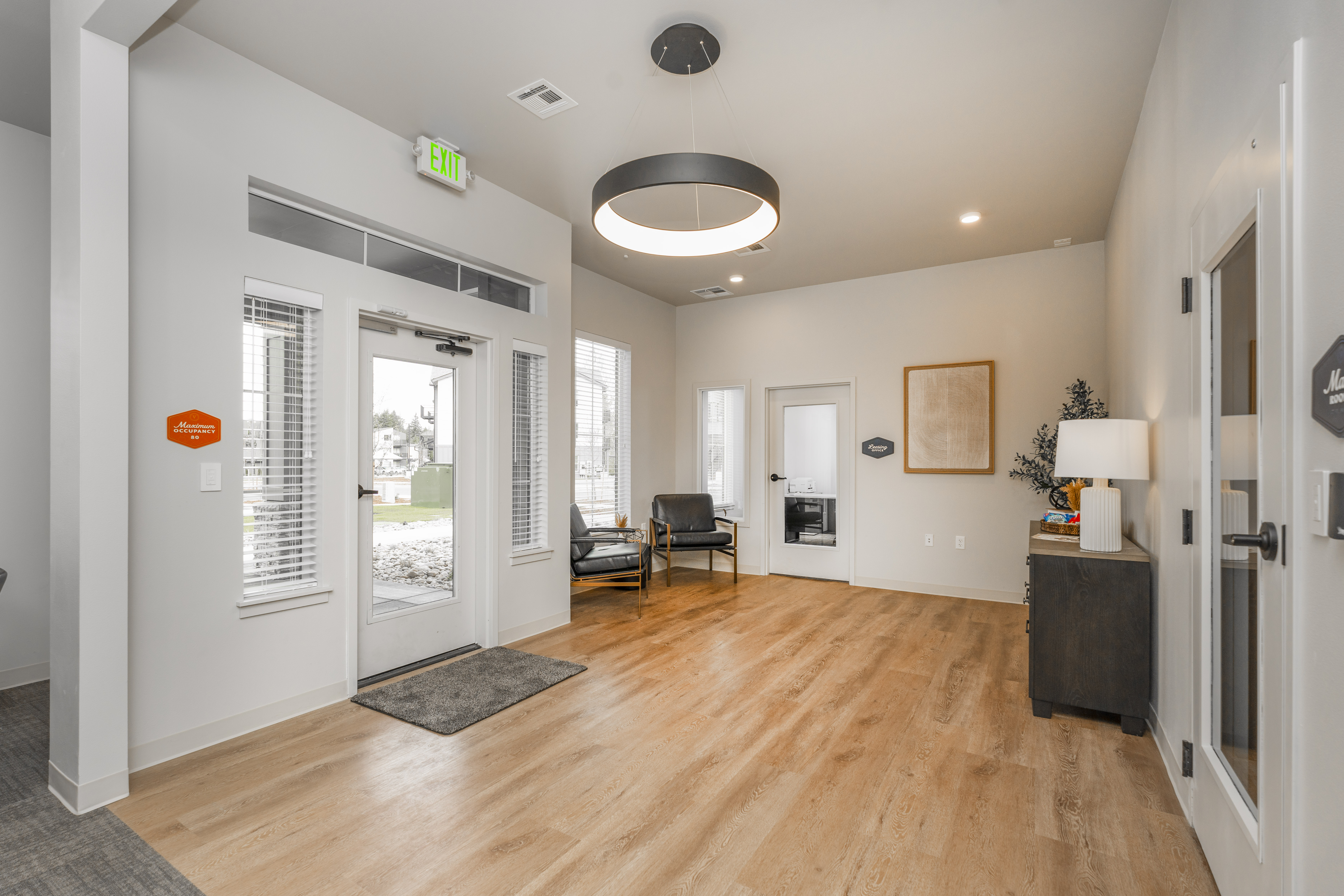 Bright modern office lobby with wood flooring, two black leather chairs by windows, a pendant light, exit door, and a sideboard with a lamp and plant.