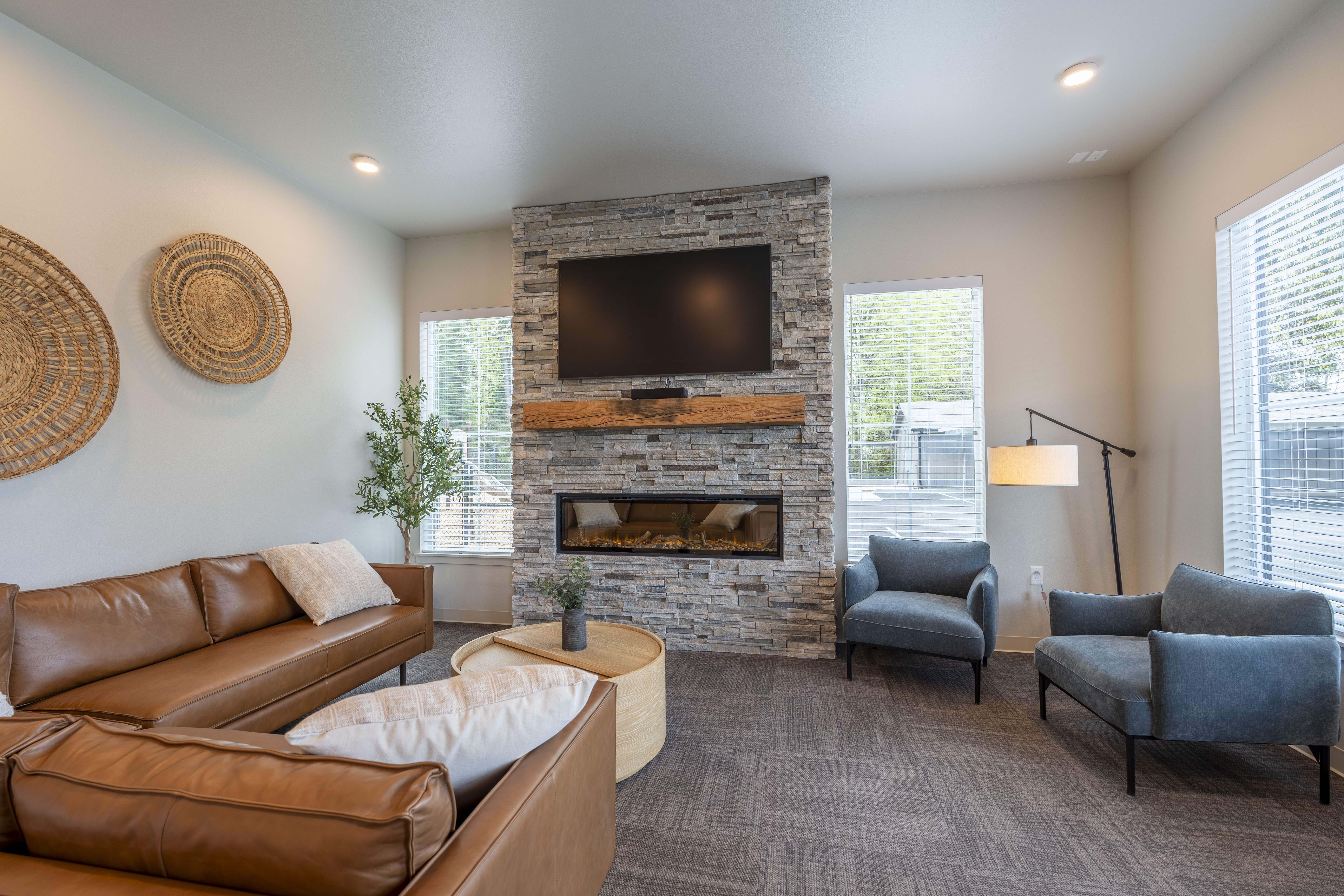Modern living room with leather sofas, two blue armchairs, a stone fireplace with a mounted TV above it, and large windows.