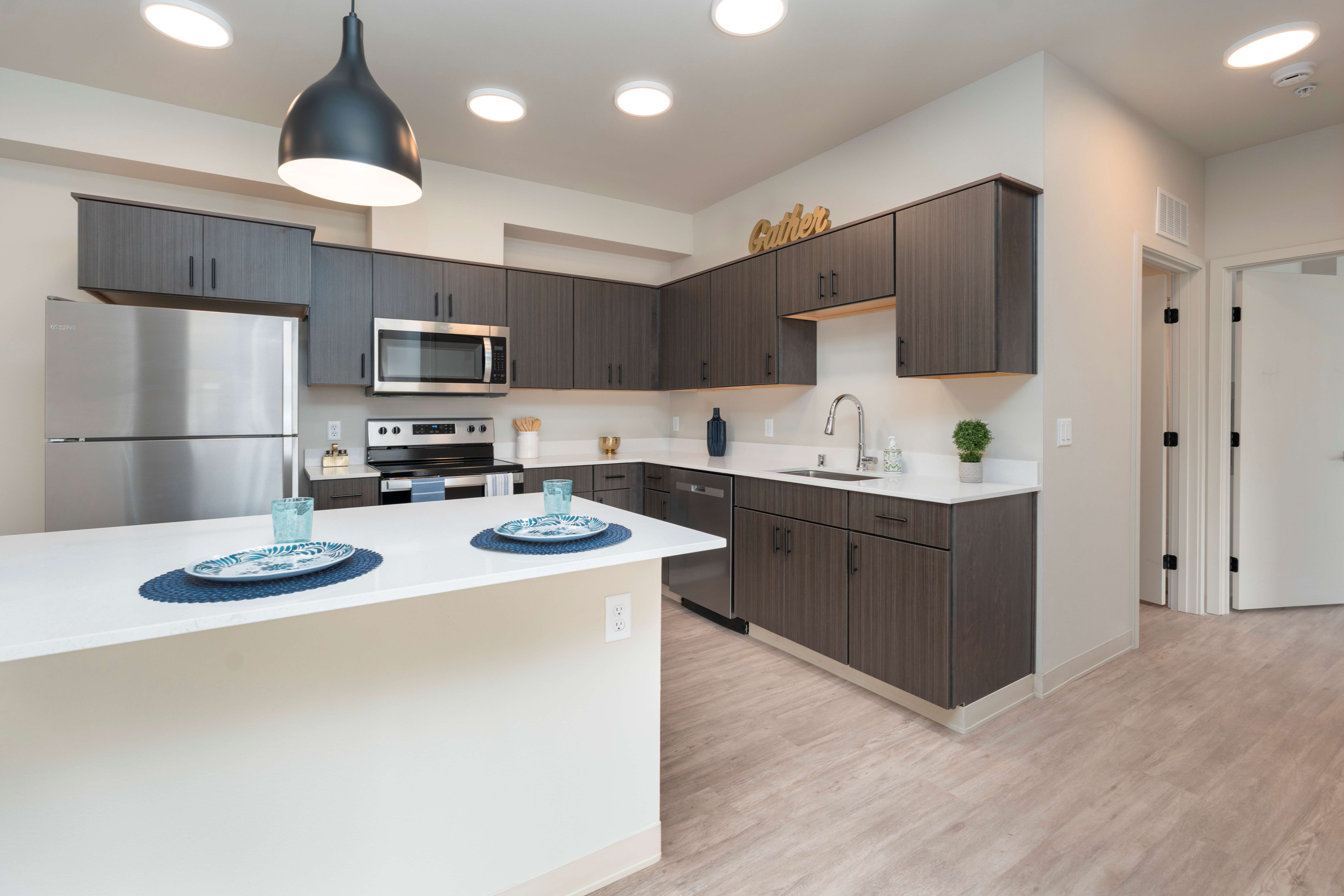 Modern kitchen with dark wood cabinets, stainless steel appliances, white countertops, and a breakfast bar set with plates and glasses.