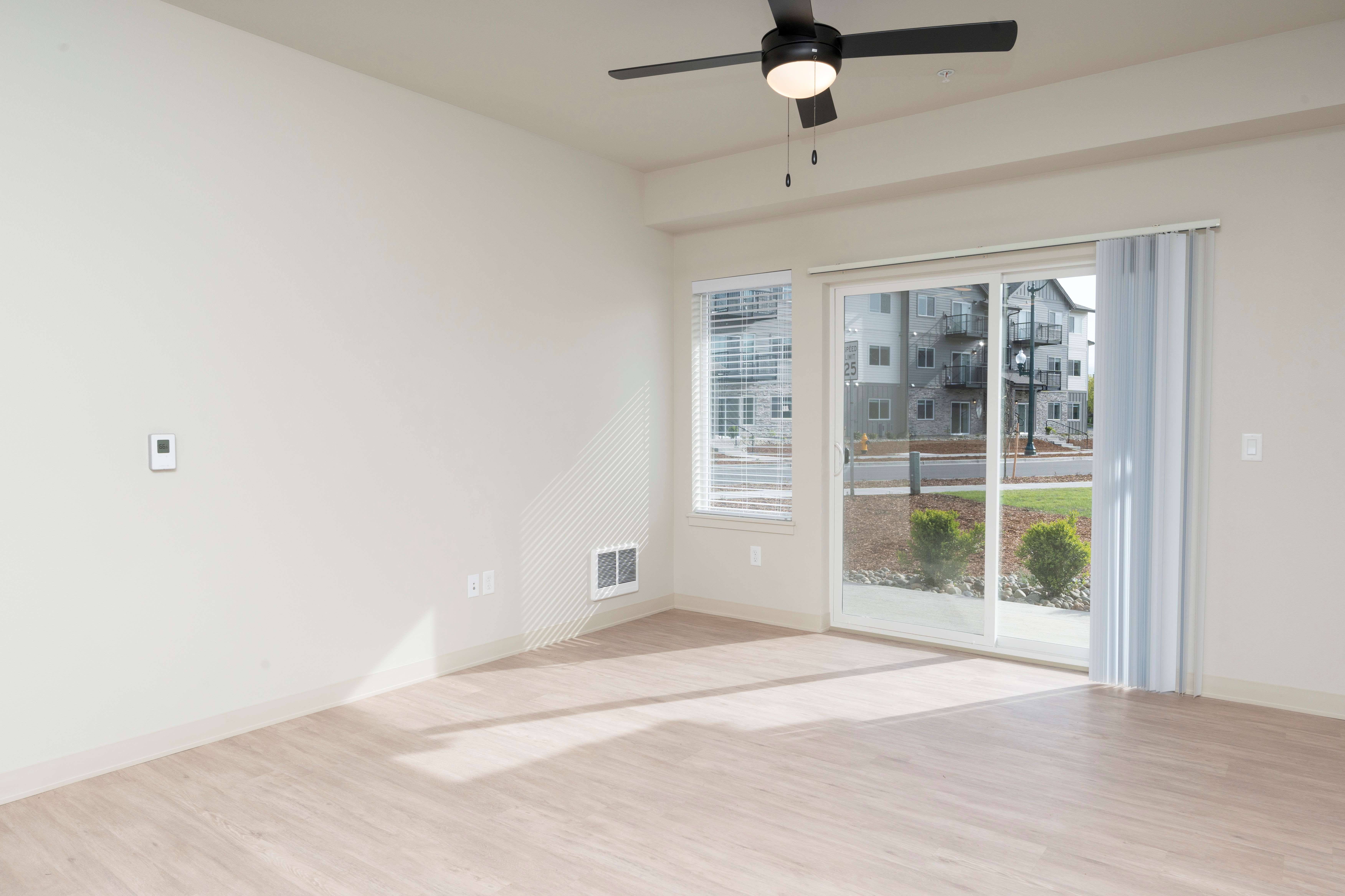Empty living room with light wood flooring, white walls, ceiling fan, and large sliding glass doors with vertical blinds.