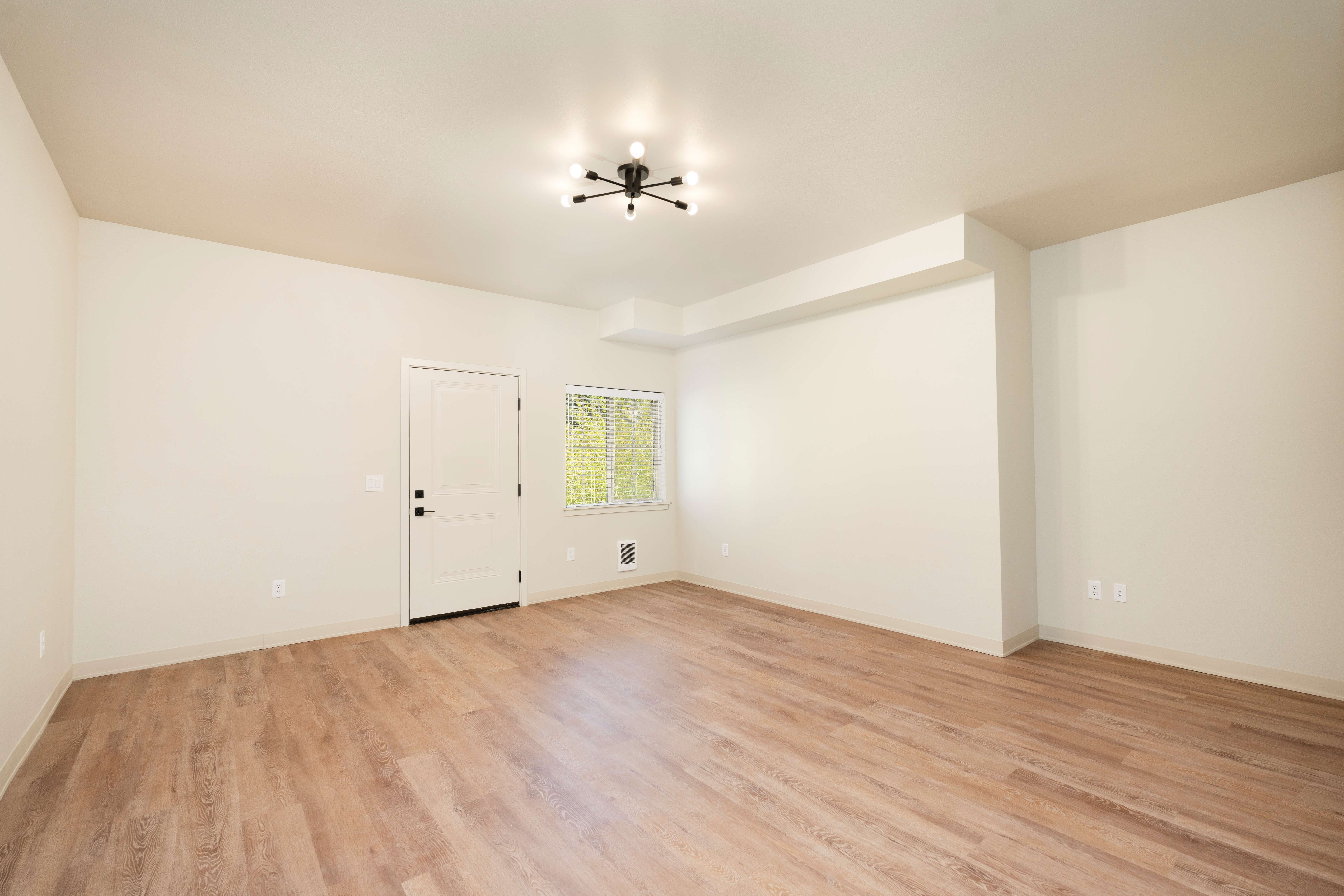 Empty room with light wooden flooring, white walls, a door, a window with blinds, and a modern ceiling light fixture.