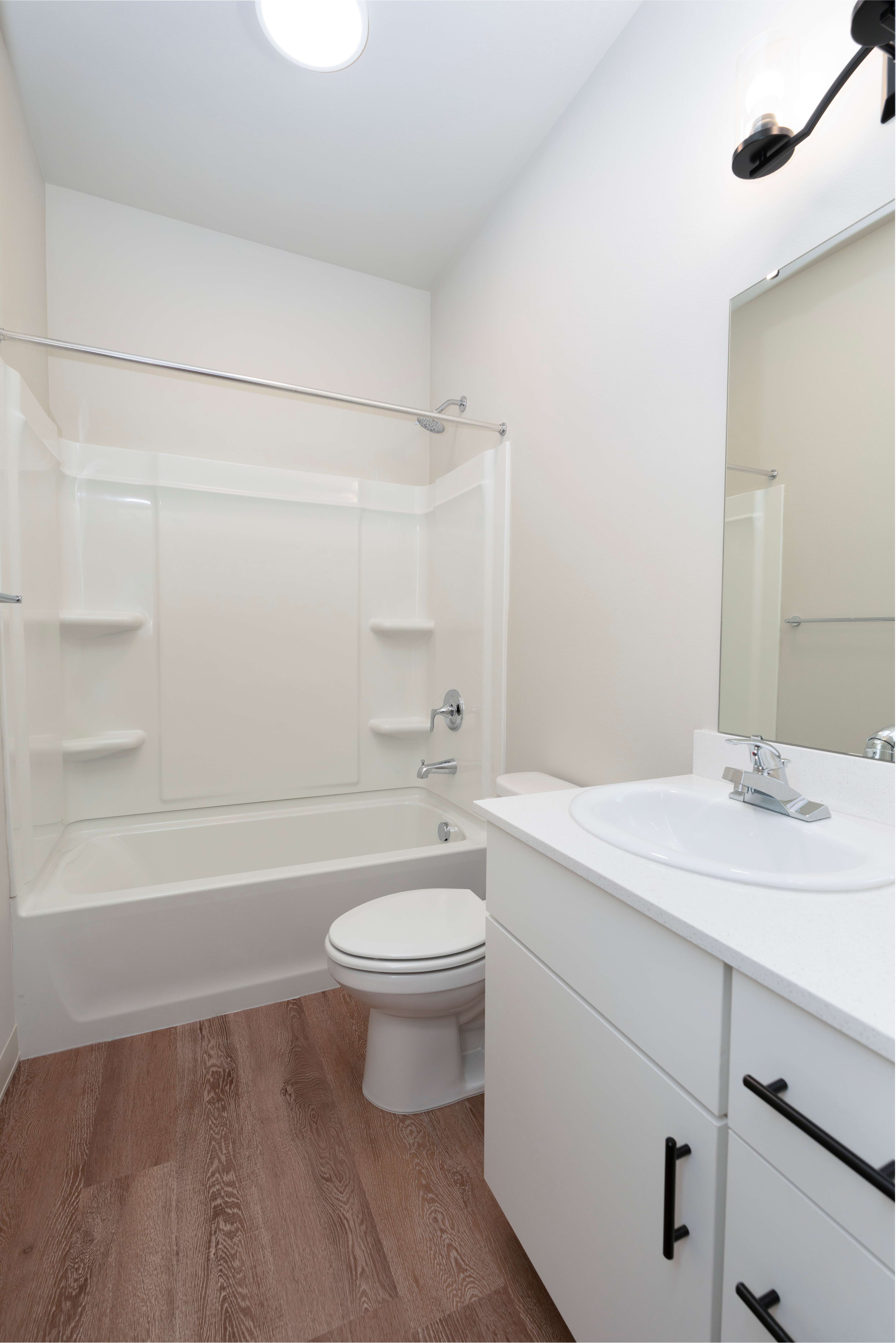 Compact bathroom with white bathtub, toilet, and sink vanity with black handles on wood floor.