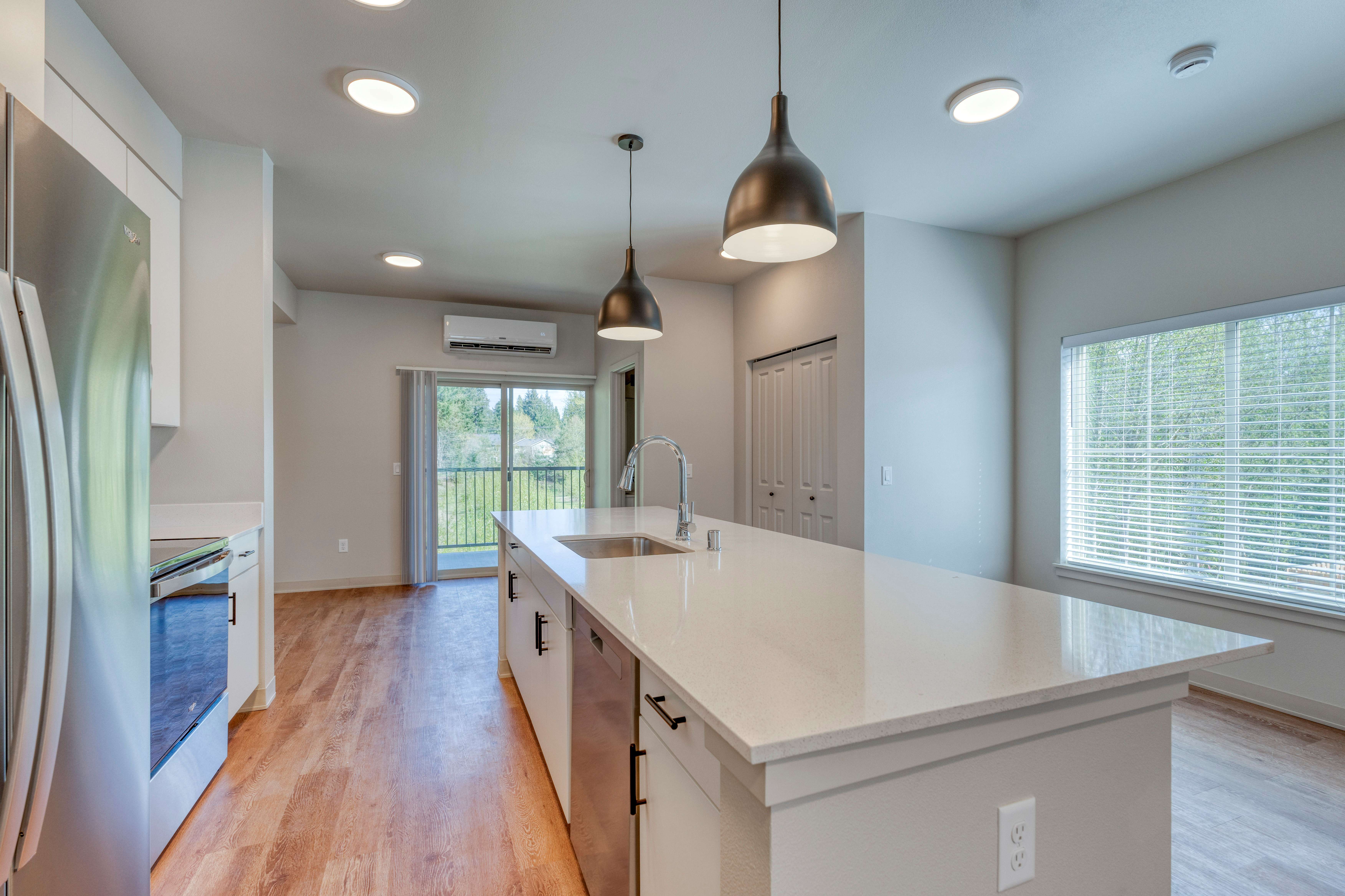 Modern kitchen with a large white island countertop, stainless steel appliances, pendant lights, and wood flooring.