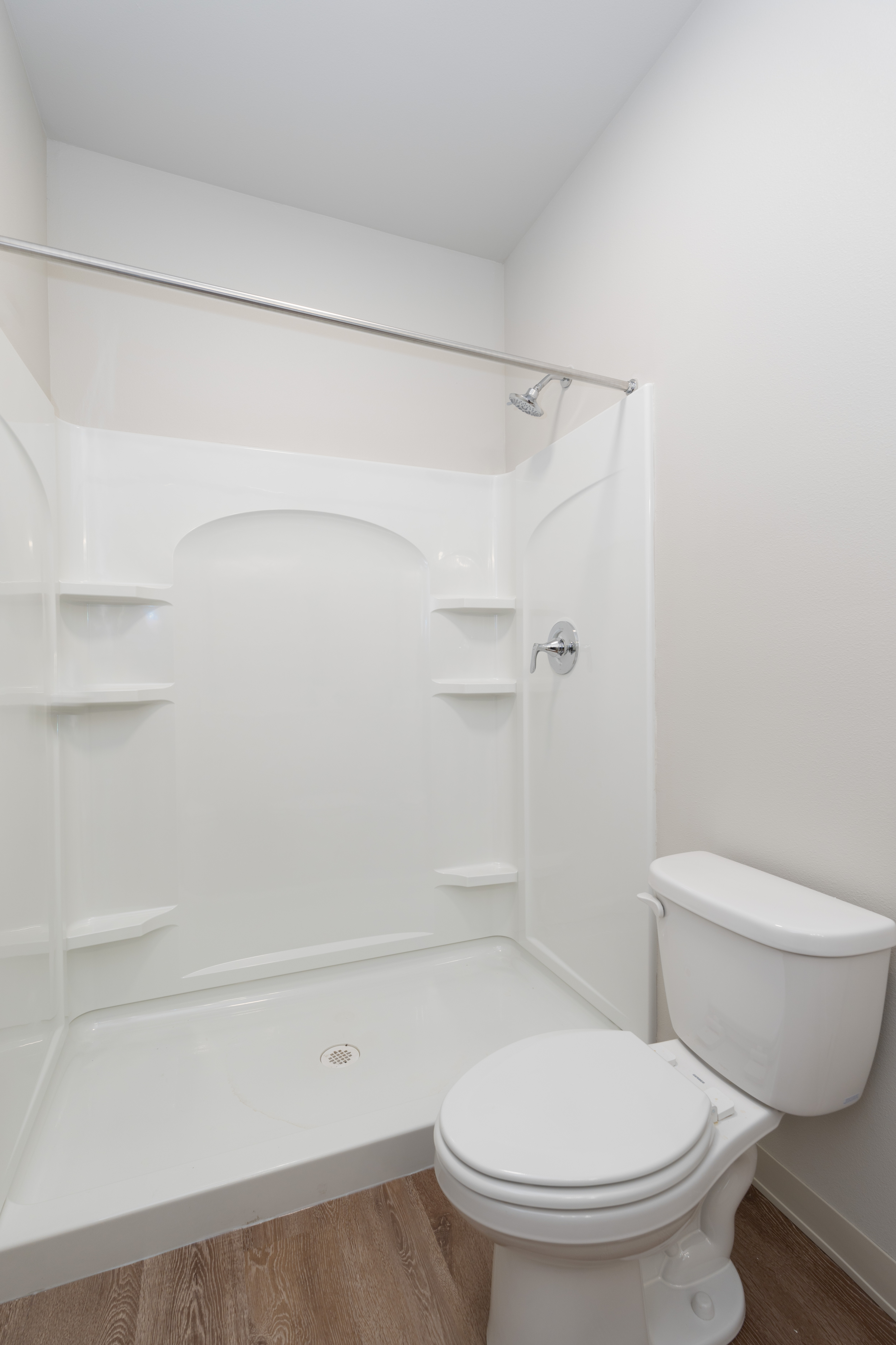 White bathroom featuring a shower with built-in shelves and a closed lid toilet on wood-like flooring.