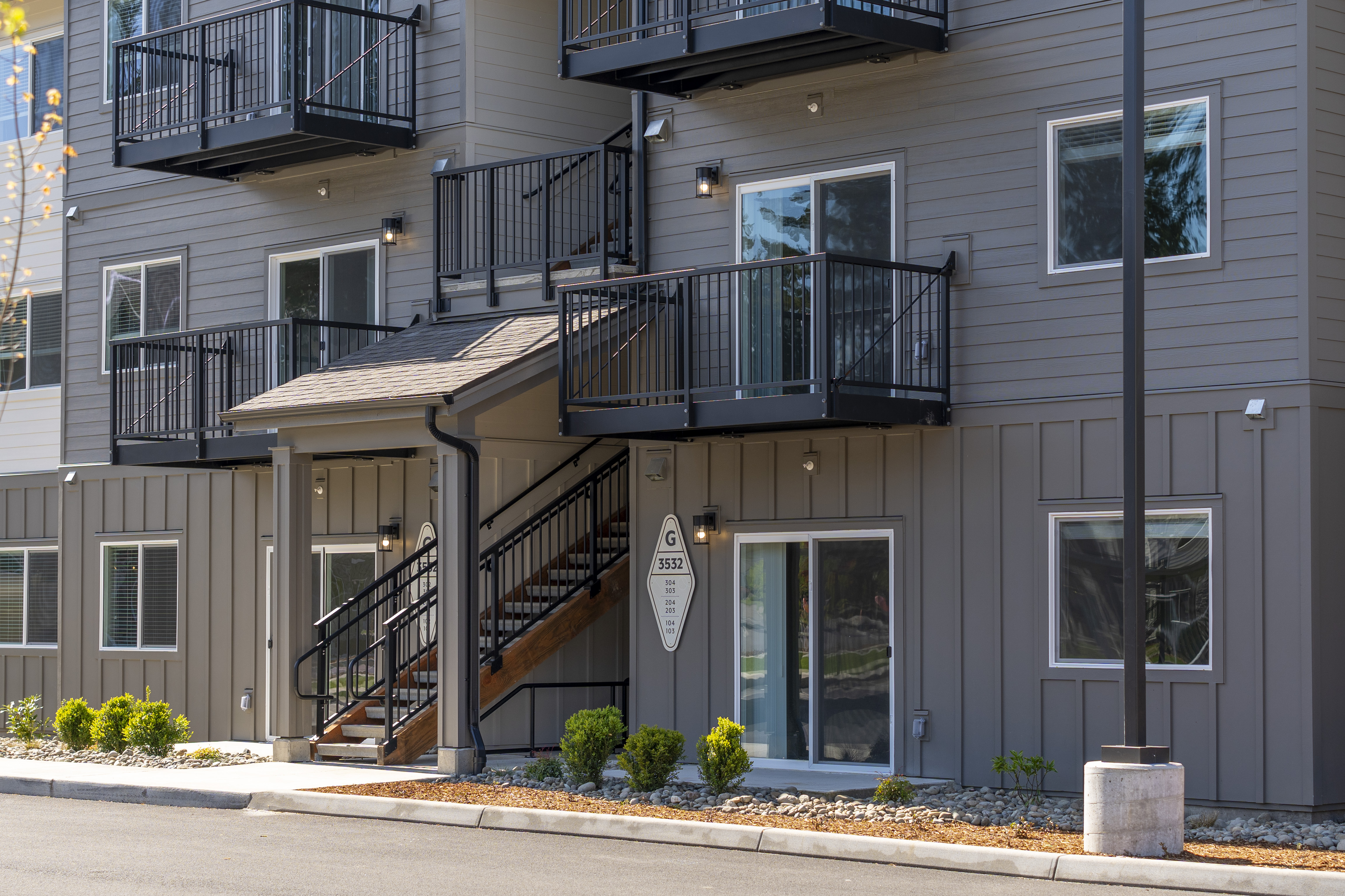 Modern multi-story apartment building exterior with balconies, stairway, sliding glass doors, and small landscaped shrubs.