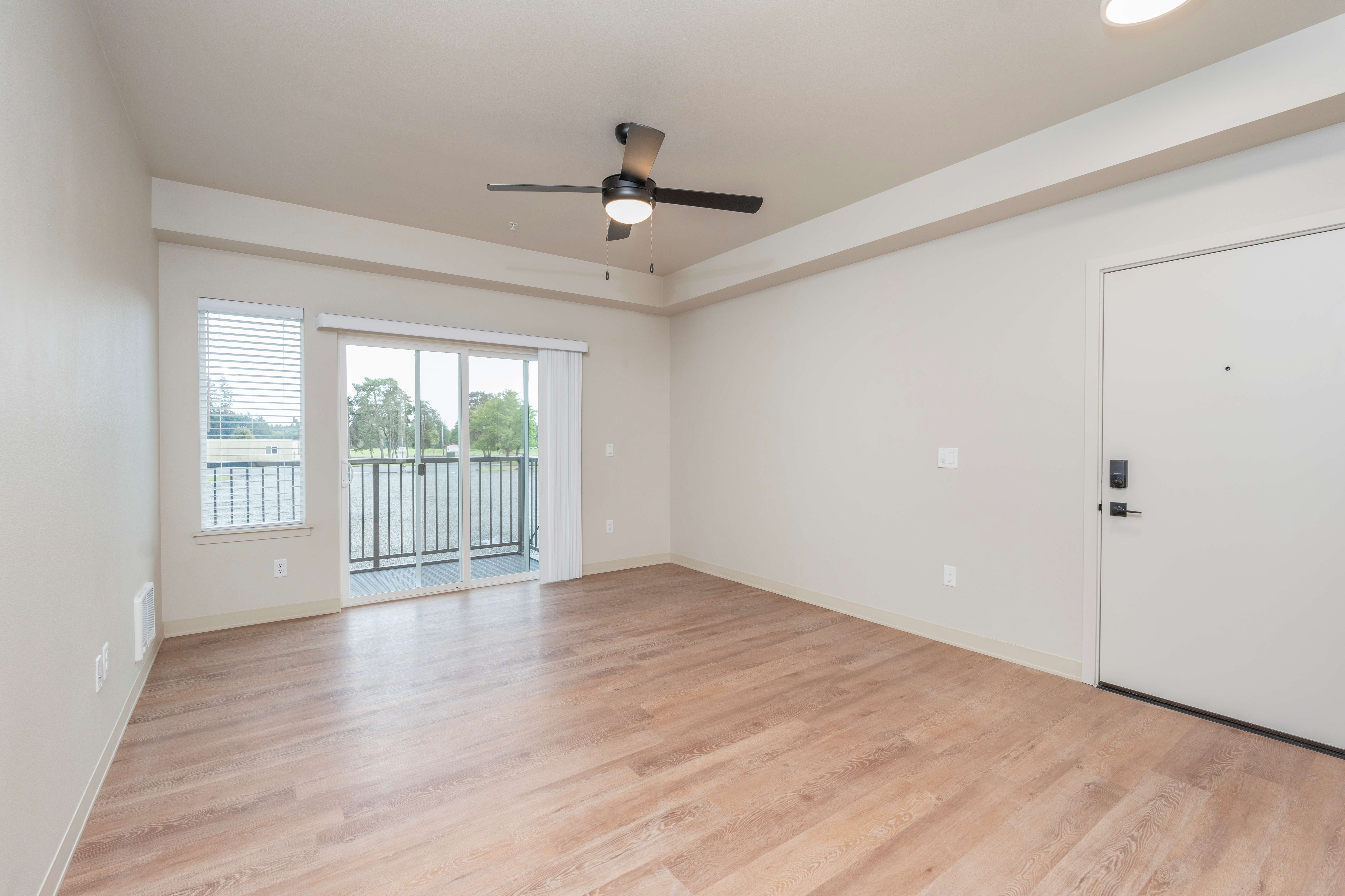 Empty living room with light wood flooring, ceiling fan, sliding glass door leading to a balcony, and white walls.