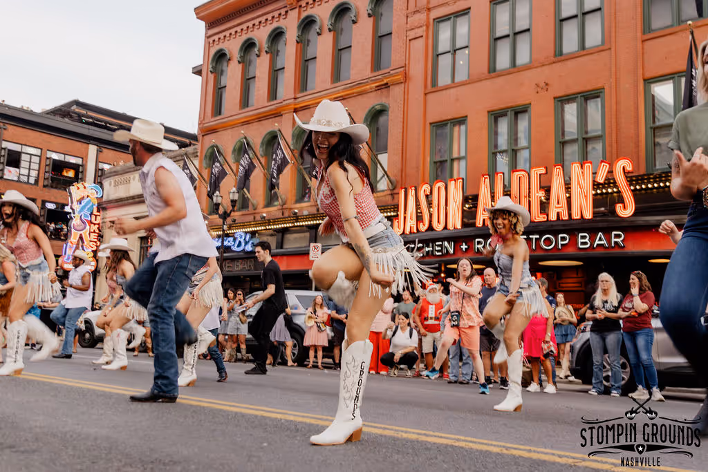Dancers in cowboy hats and fringed outfits performing on a street with a crowd and Jason Aldean's bar in the background.