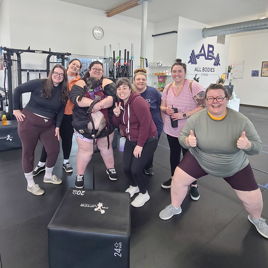 A group of individuals surround coach Christina and coach Koko after a powerlifting class