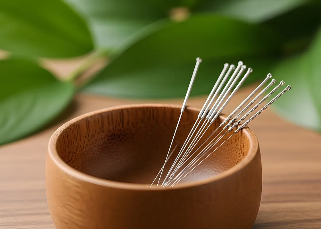 Thin stainless steel acupuncture needles arranged inside a small wooden bowl on a wooden surface with green leaves blurred in the background.