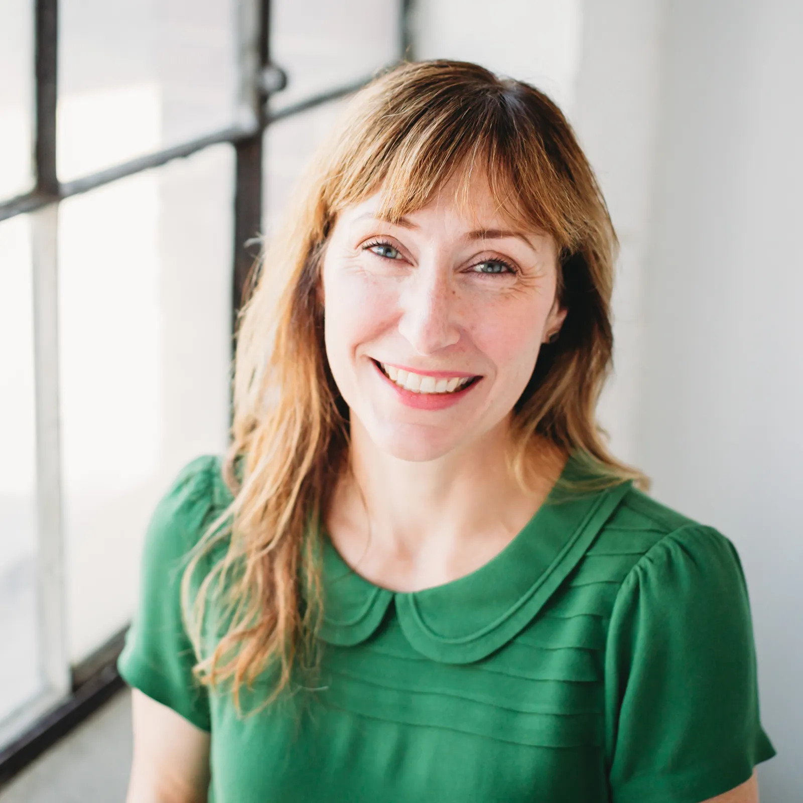 Smiling woman with light brown hair wearing a green top with a Peter Pan collar, standing by a window.
