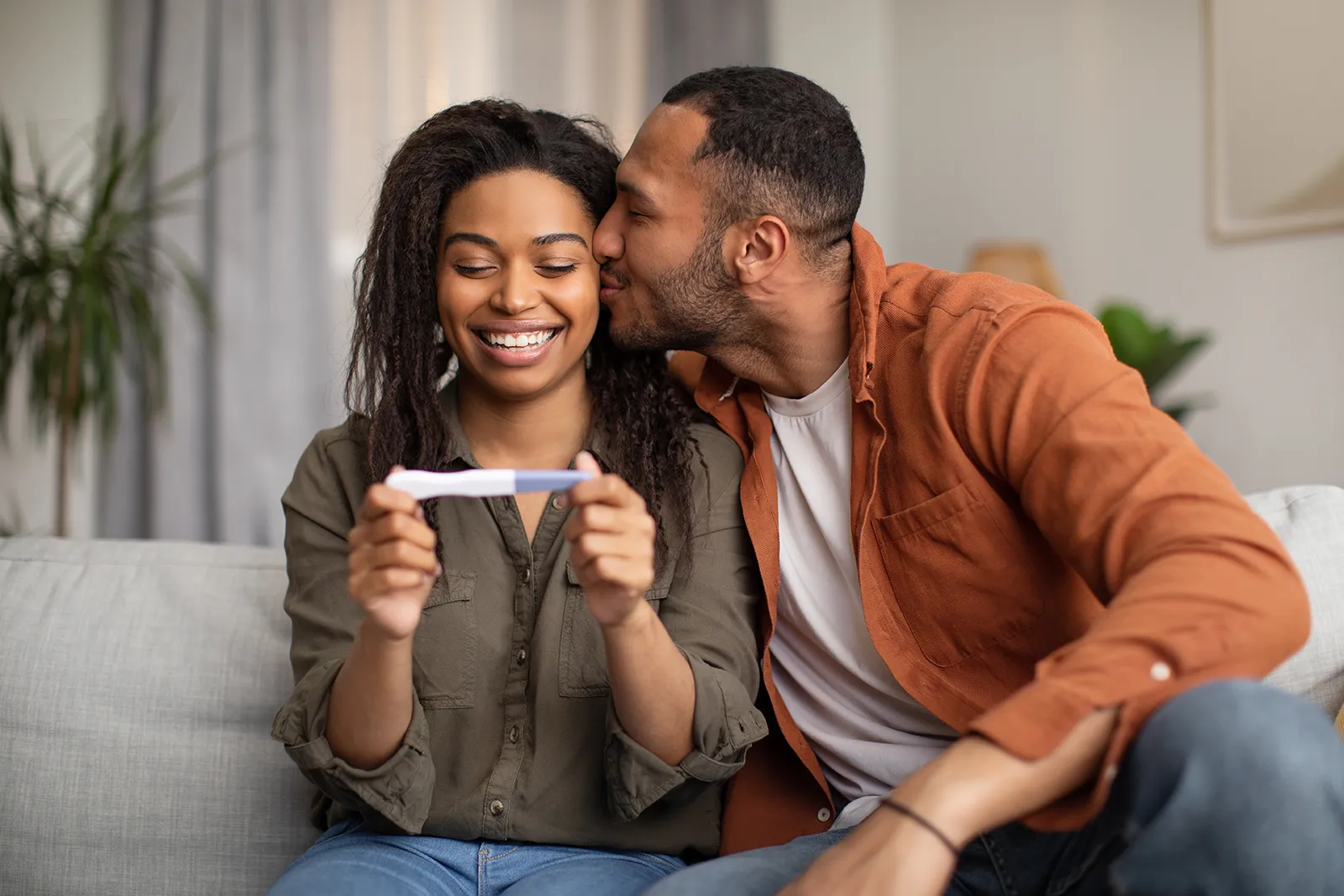 Smiling woman holding a positive pregnancy test while a man kisses her cheek on a couch.