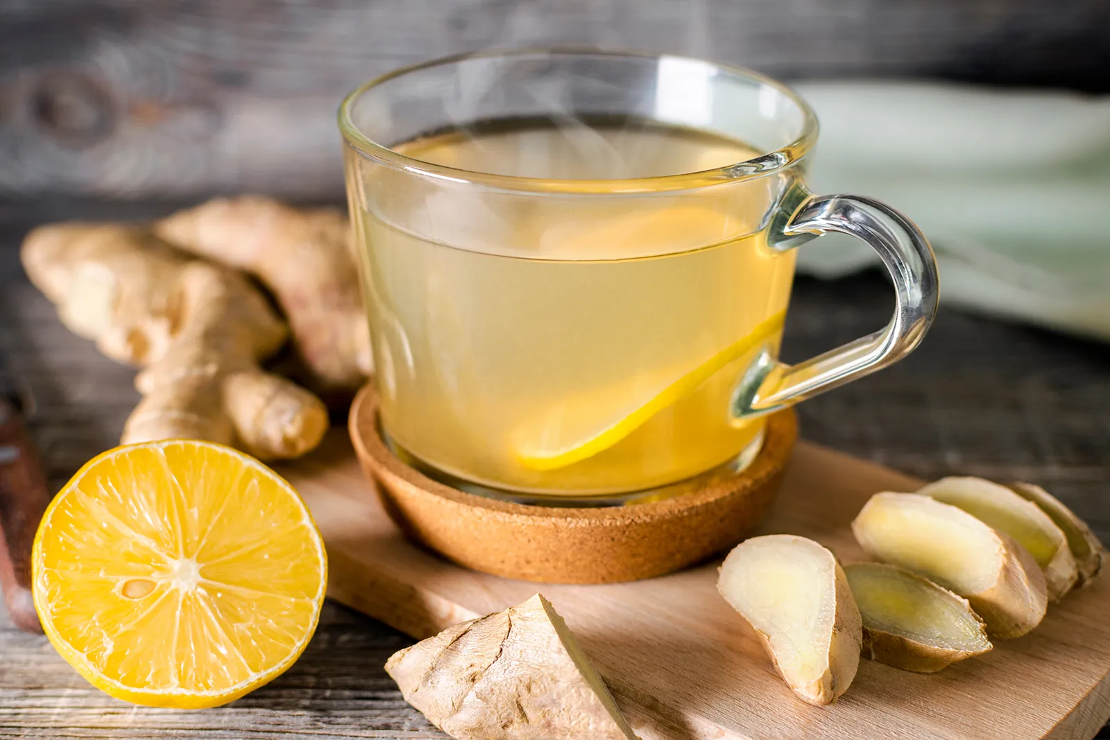 Glass cup of steaming ginger tea with lemon slice, fresh ginger roots, and lemon half on a wooden surface.