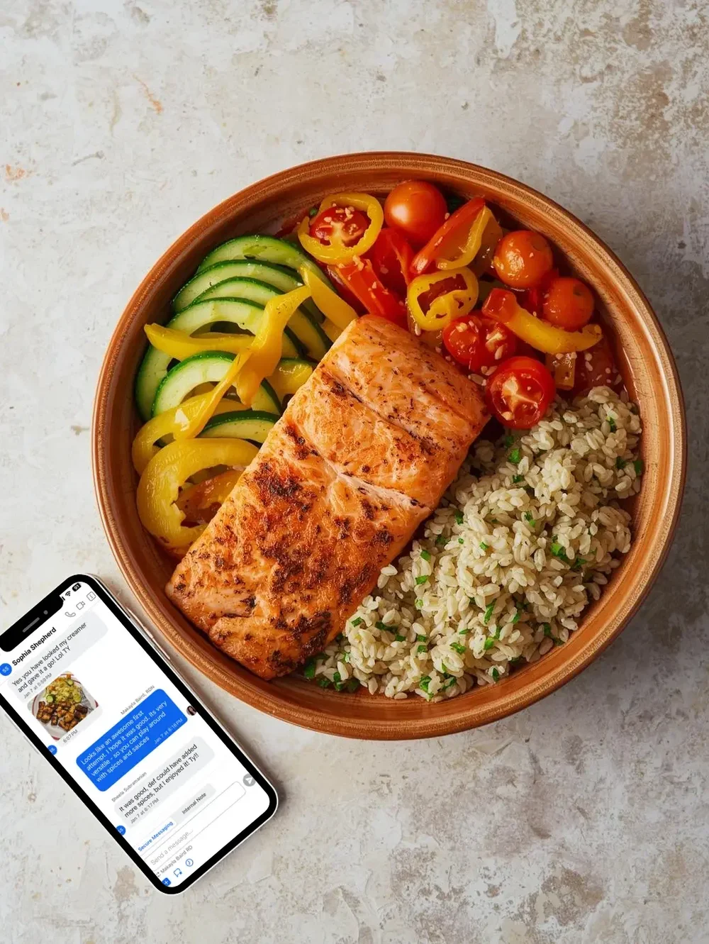 Grilled salmon with yellow bell peppers, cherry tomatoes, zucchini slices, and barley in a round bowl next to a smartphone showing a text conversation.