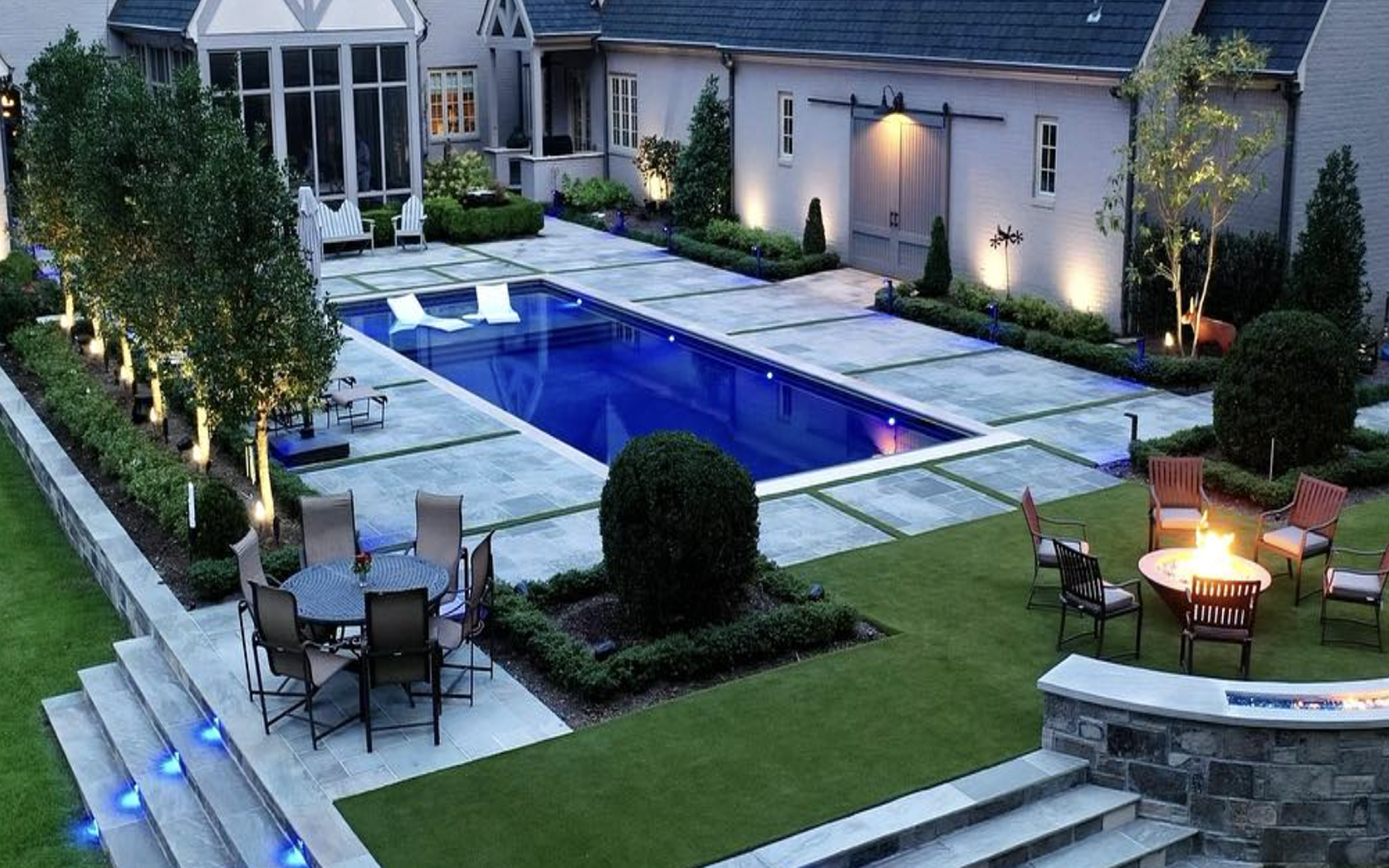 Evening view of a modern backyard with a lit rectangular pool, seating areas, fire pit, and trees.