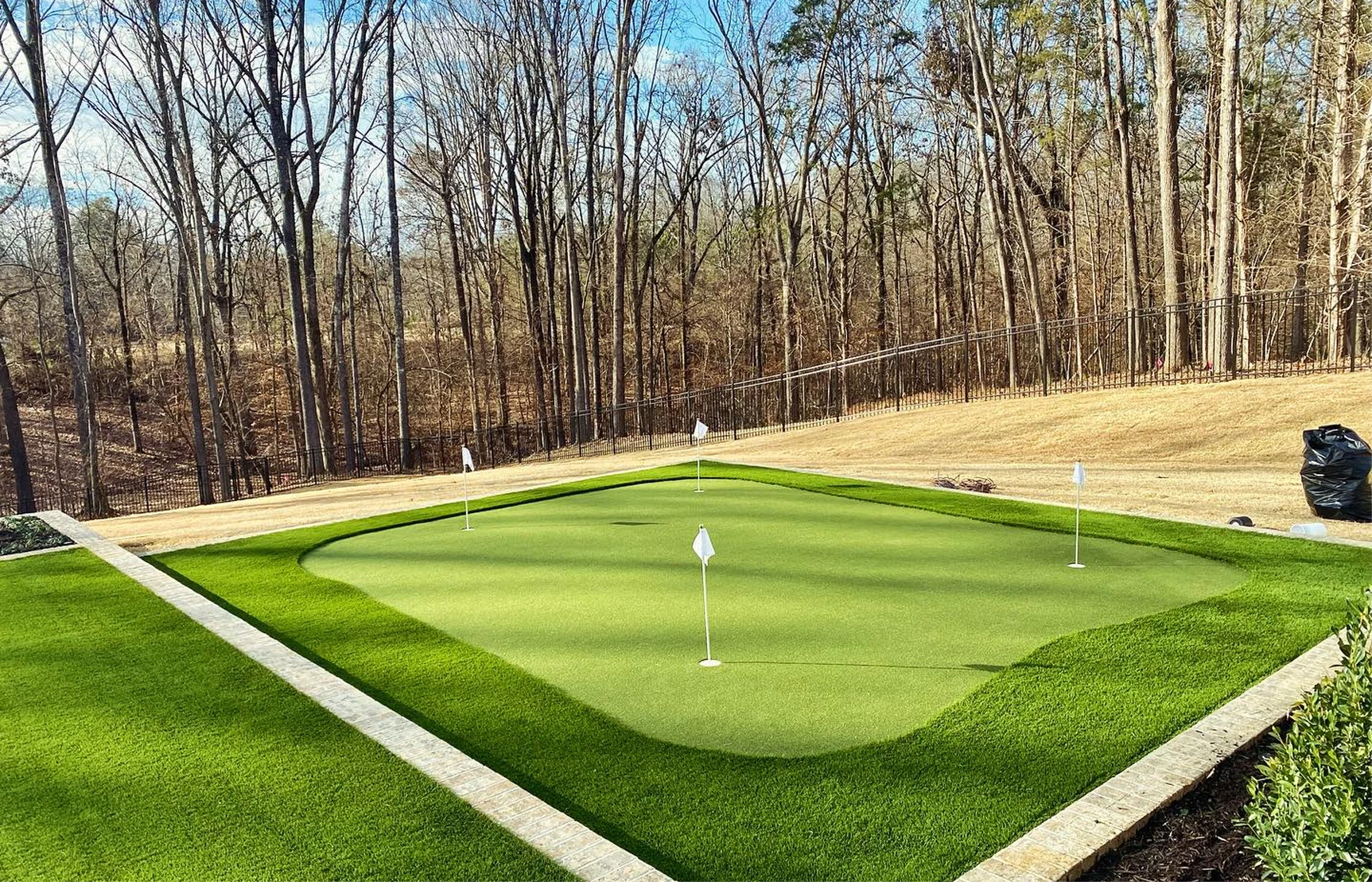 A backyard golf putting green with four white flags surrounded by trees and lawn.