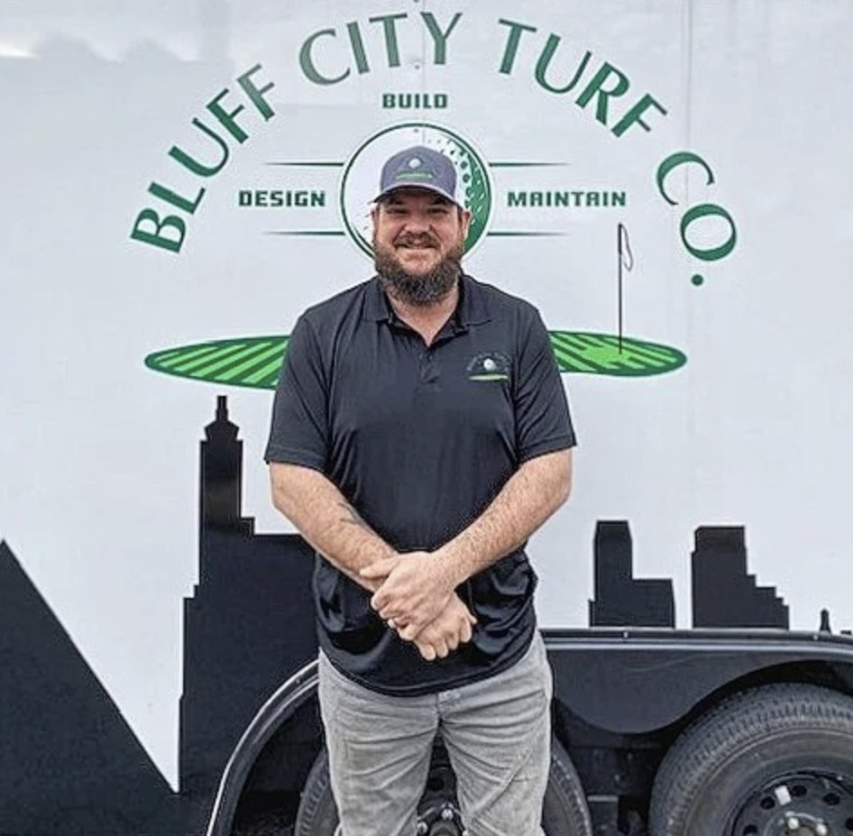 Man in a black polo and cap stands smiling in front of a Bluff City Turf Co. logo with city skyline.