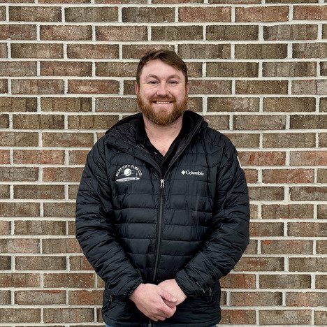 Man with beard wearing a black Columbia jacket standing and smiling against a brick wall.