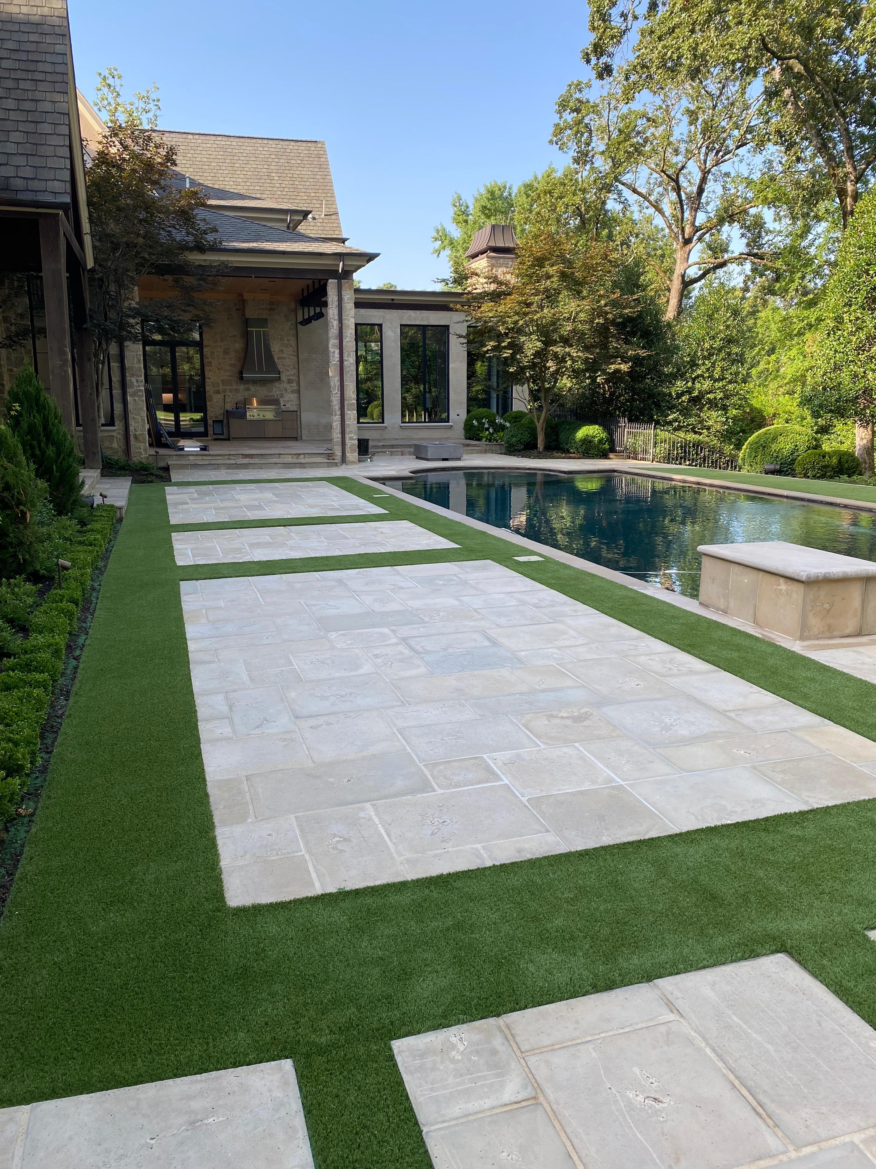Residential backyard with stone patio, artificial grass strips, a pool, and mature trees under blue sky.