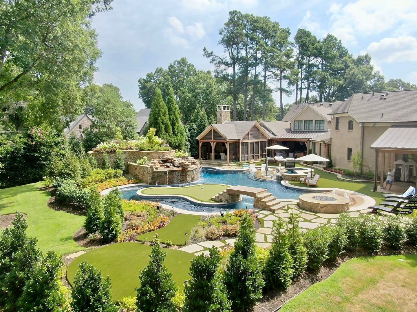 Luxury backyard with pool, waterfall, fire pit, putting green, lounge chairs, and lush landscaping.