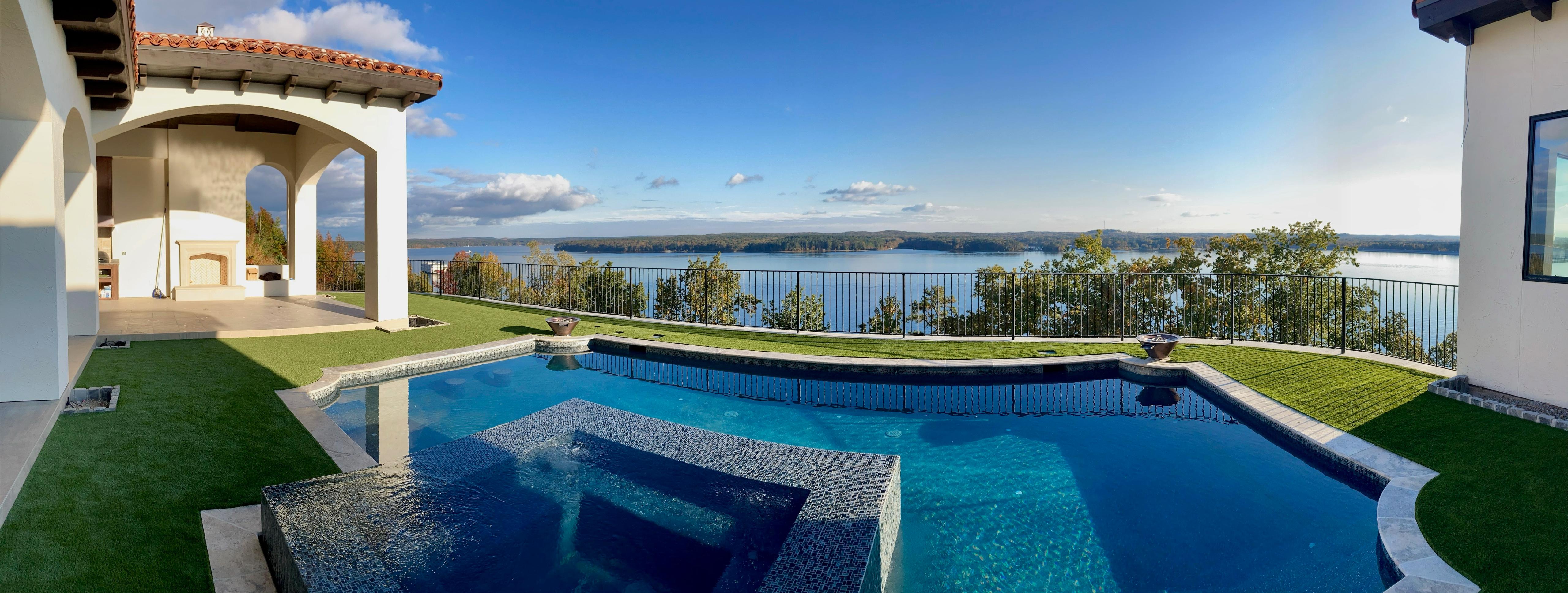 Modern house patio with pool and hot tub overlooking a lake with clear blue sky.