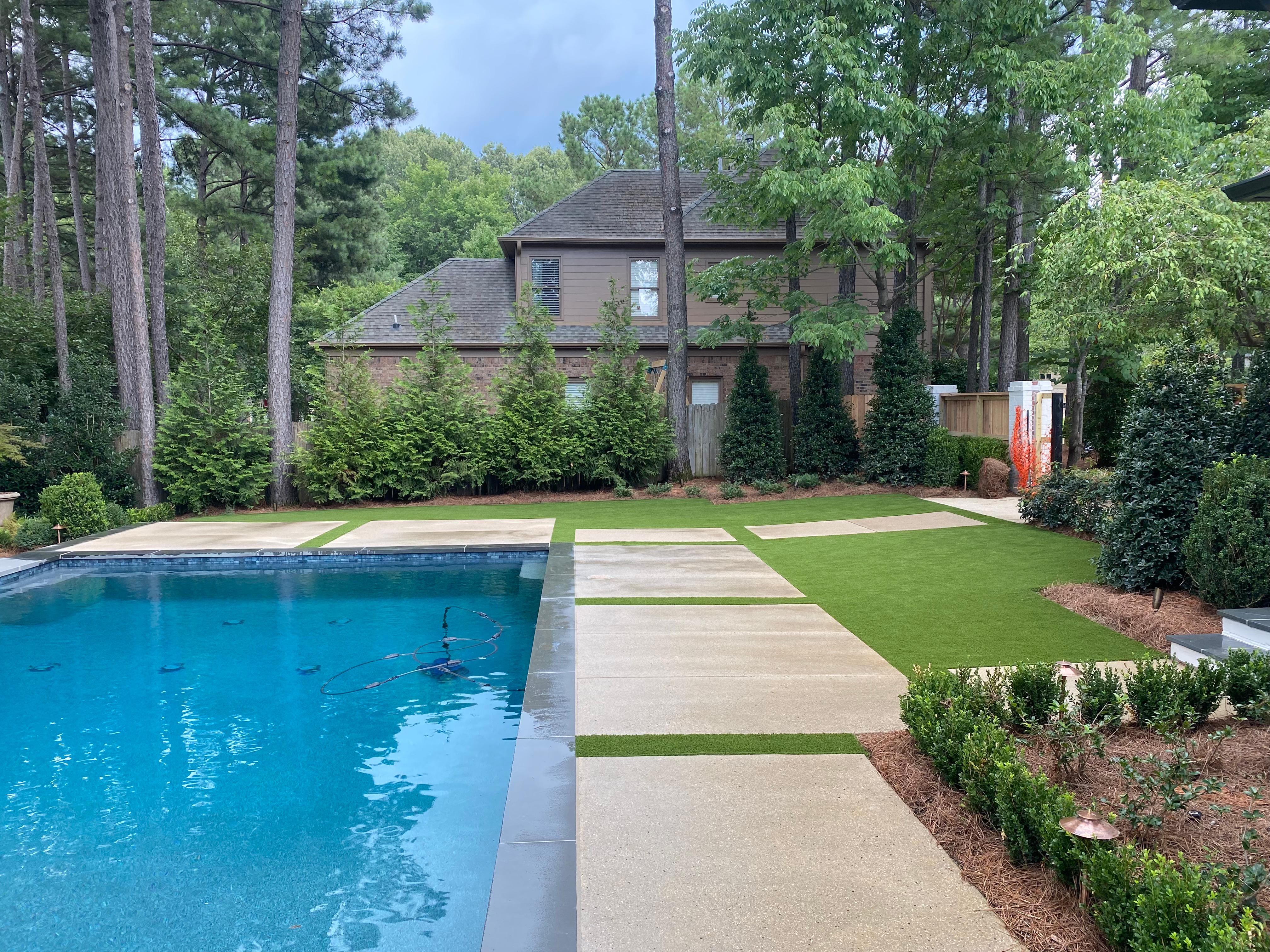 Backyard with a blue swimming pool, concrete patio, green lawn, and surrounding trees and shrubs.