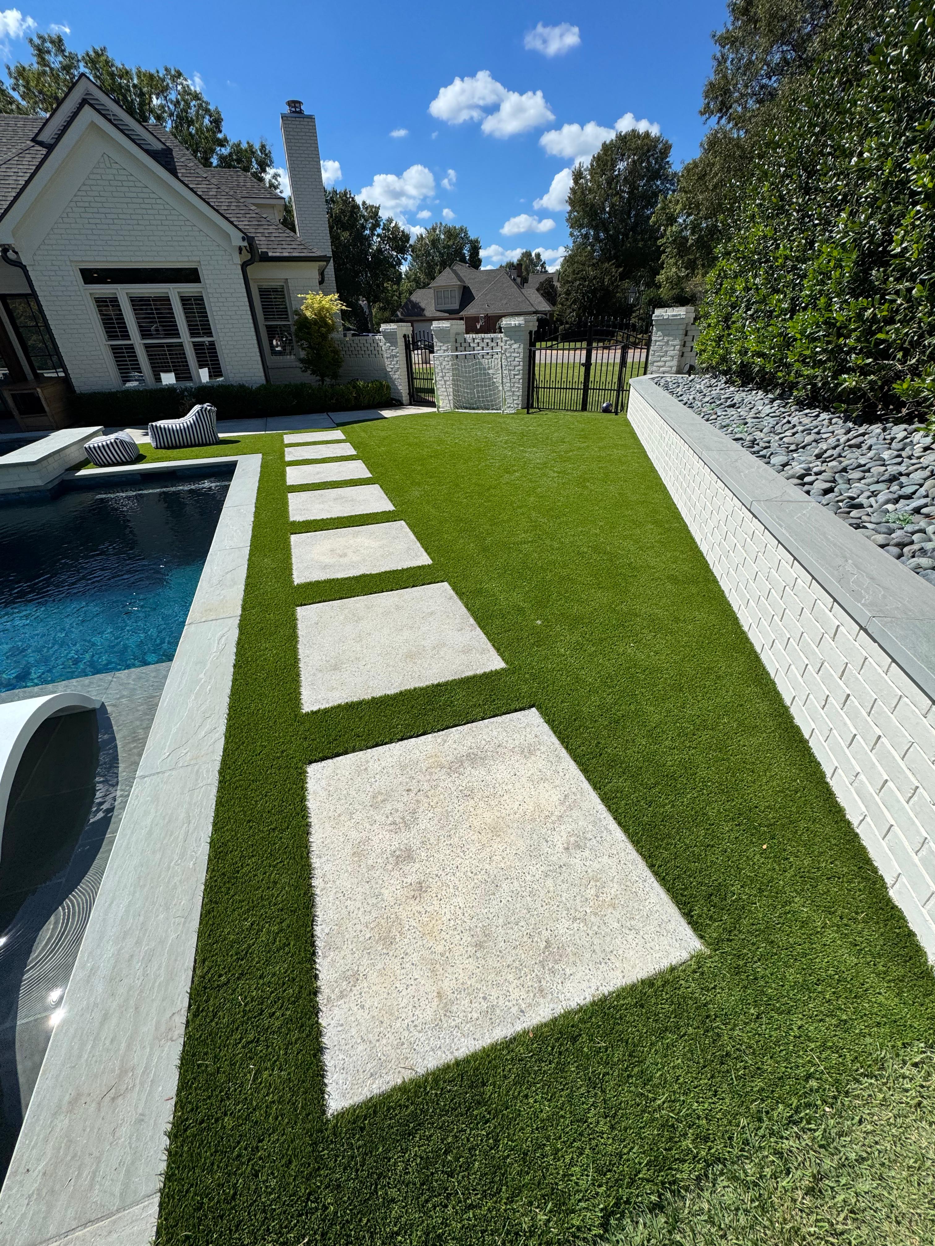 Backyard with swimming pool, stepping stones on green lawn, goal net by white brick wall and black gate.