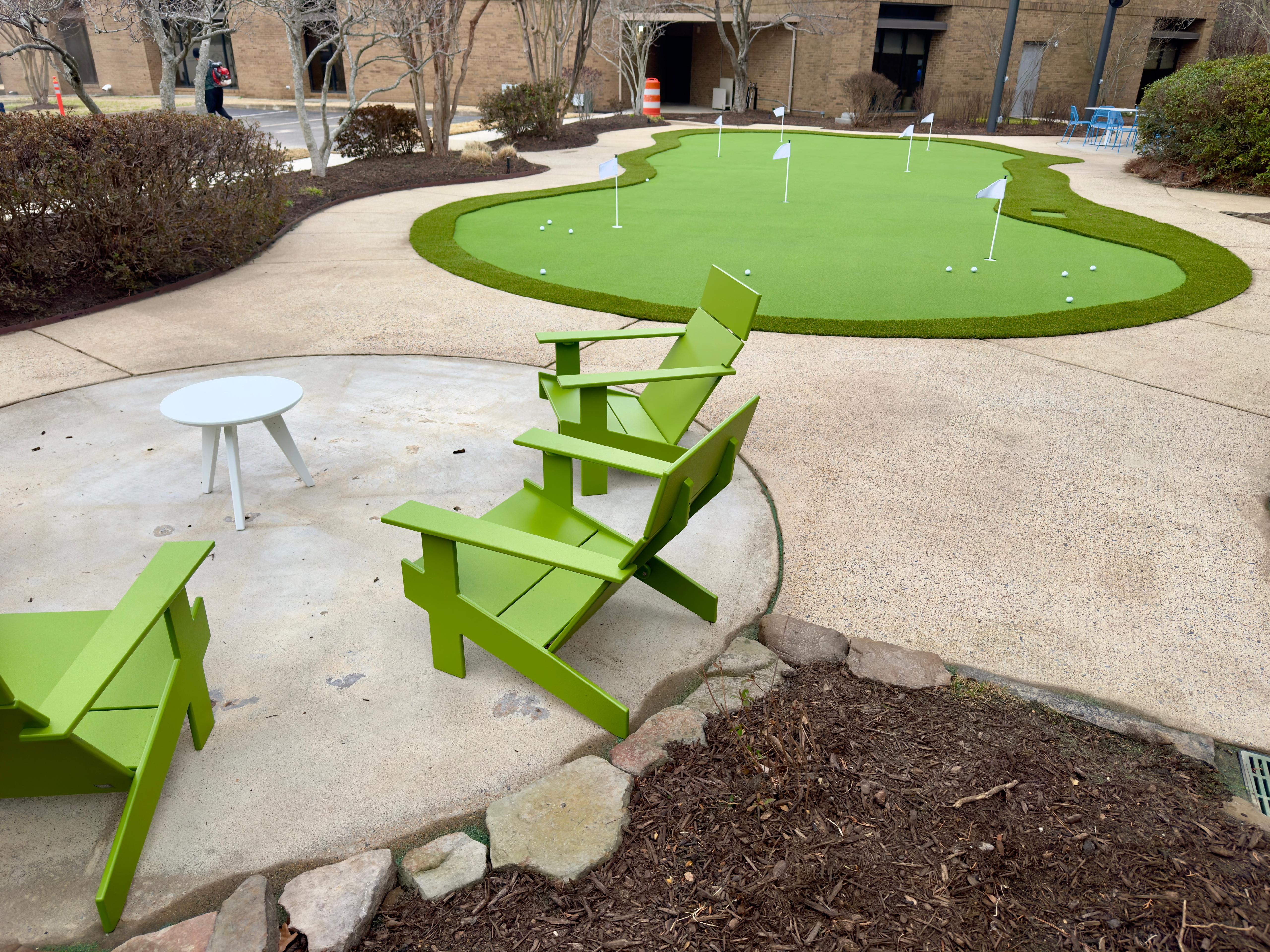 Outdoor green chairs and small table near a putting green with golf balls and flags.