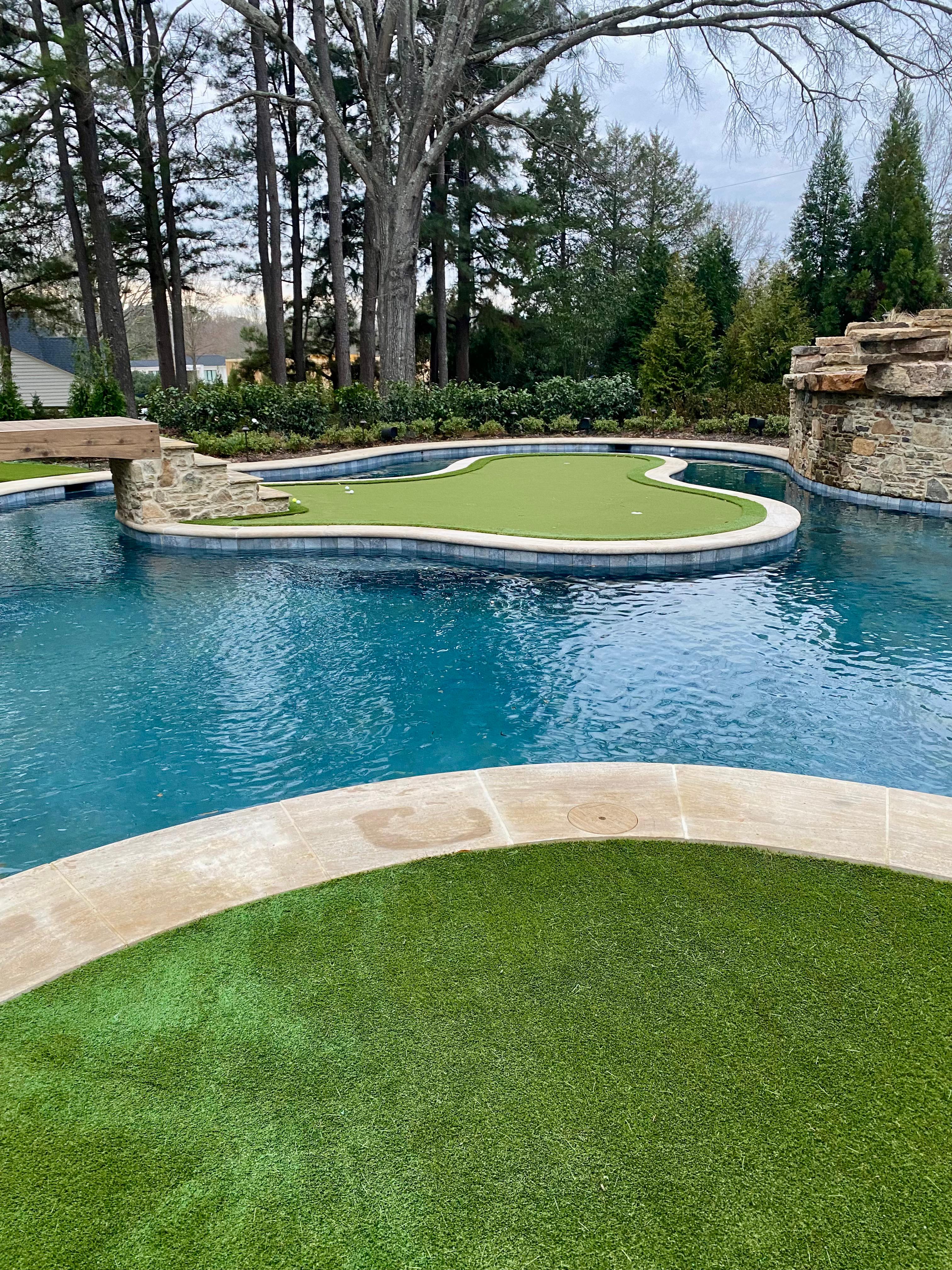 Swimming pool with a putting green island and stone steps, surrounded by trees and greenery.