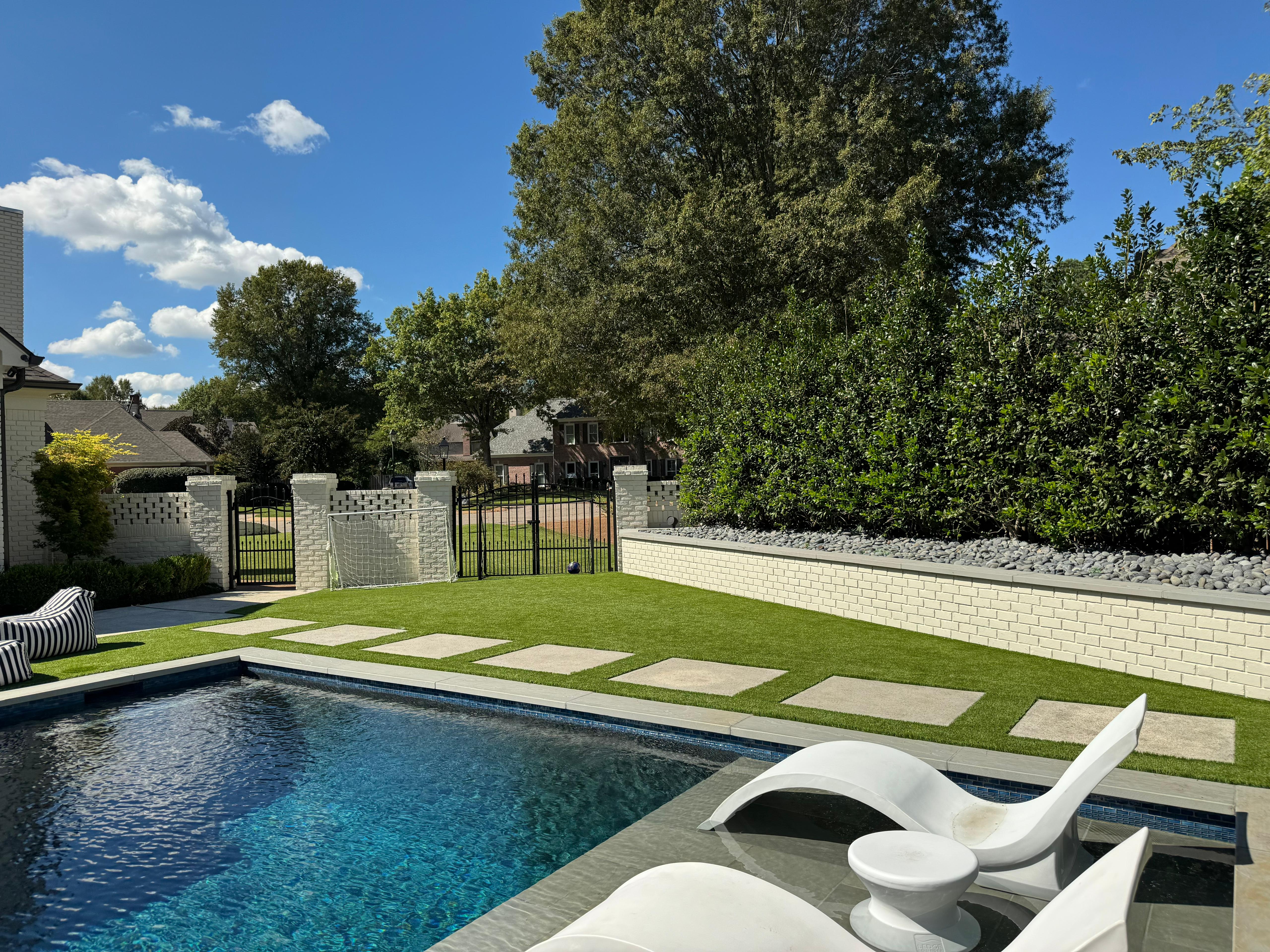 Backyard pool with two white lounge chairs in shallow water, green lawn, and trees under a blue sky.