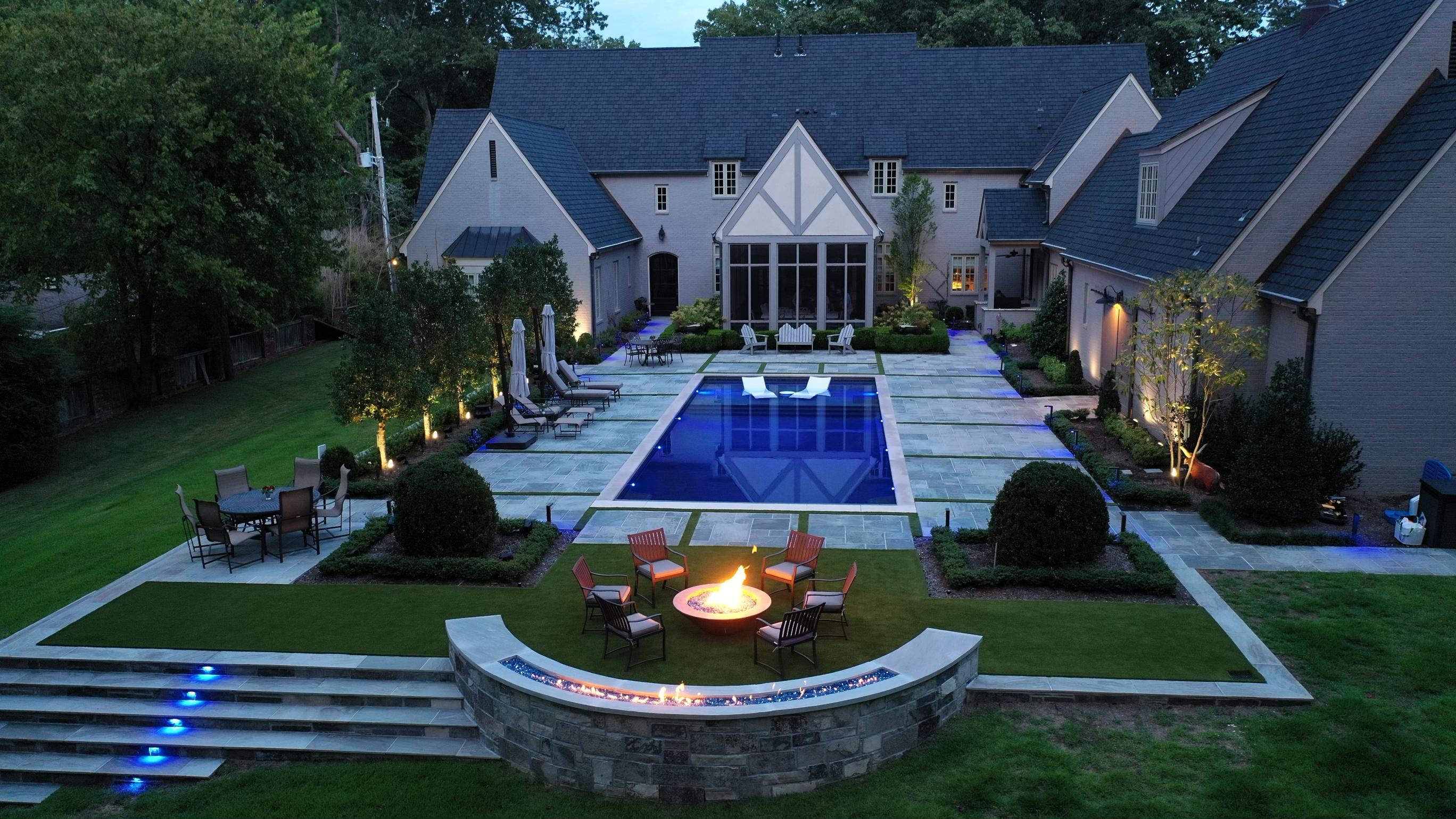 Backyard with modern house, illuminated pool, outdoor seating, fire pit, and landscaped garden at dusk.