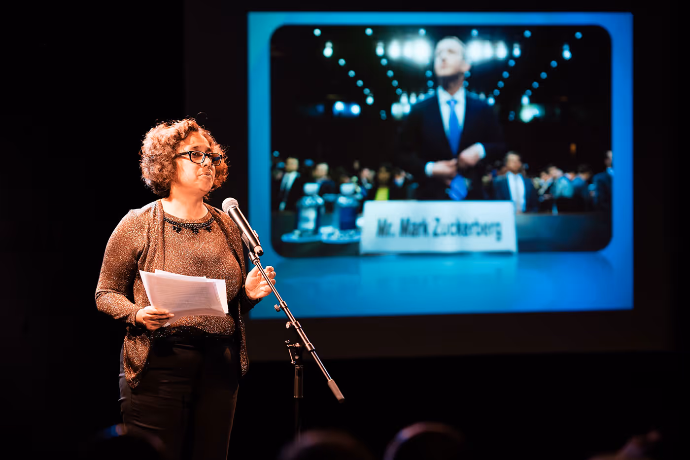 Woman with glasses speaking into a microphone on stage holding papers with a blurred projected image of Mark Zuckerberg behind her.