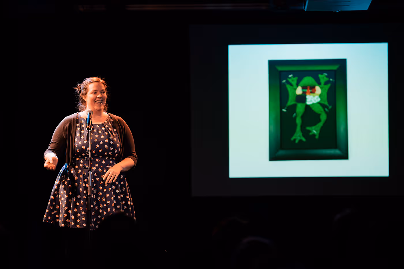 Woman in a patterned dress and cardigan speaking on stage next to a screen displaying a framed diagram of a frog.