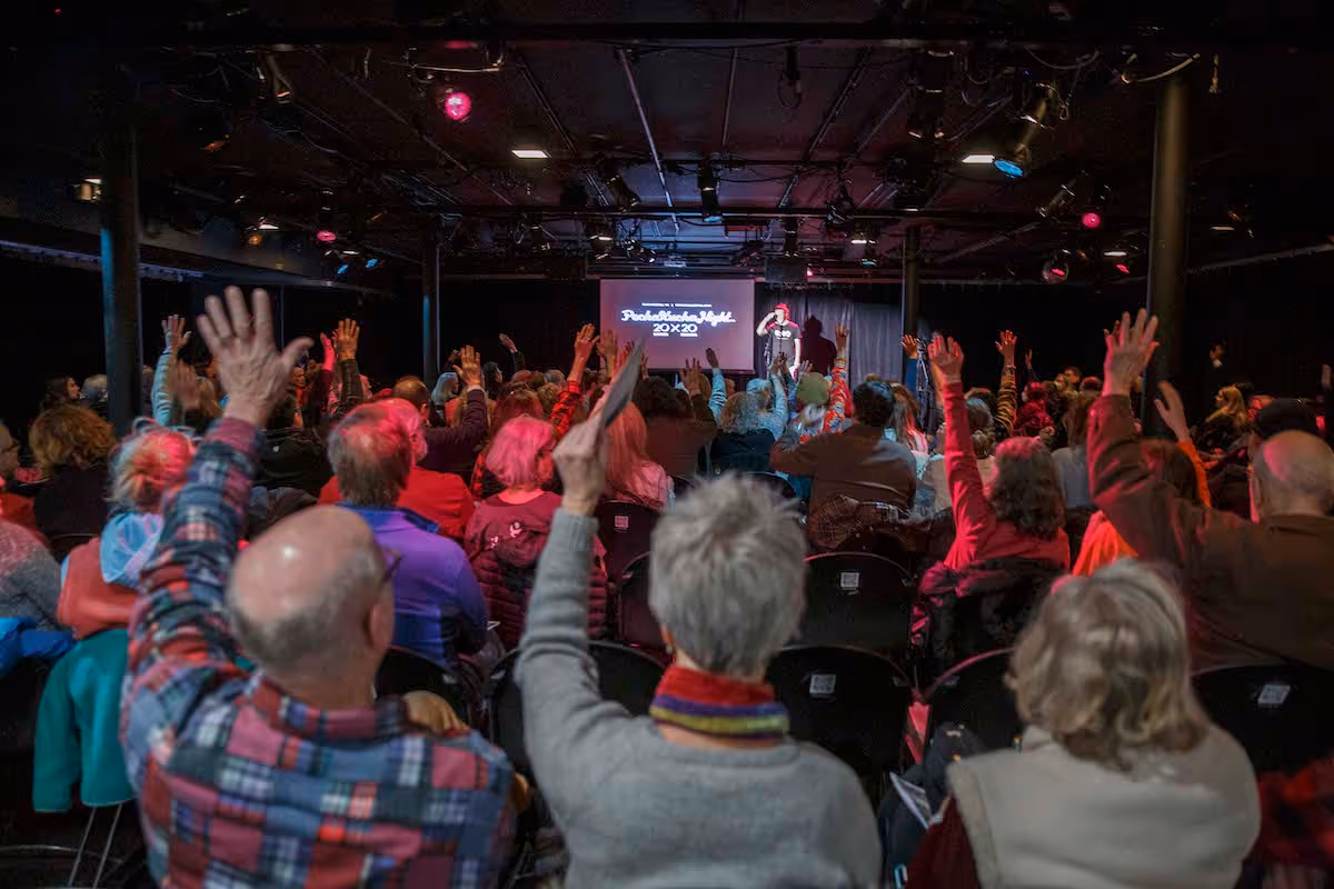 Audience in a dimly lit theater raising their hands during a PechaKucha Night event.