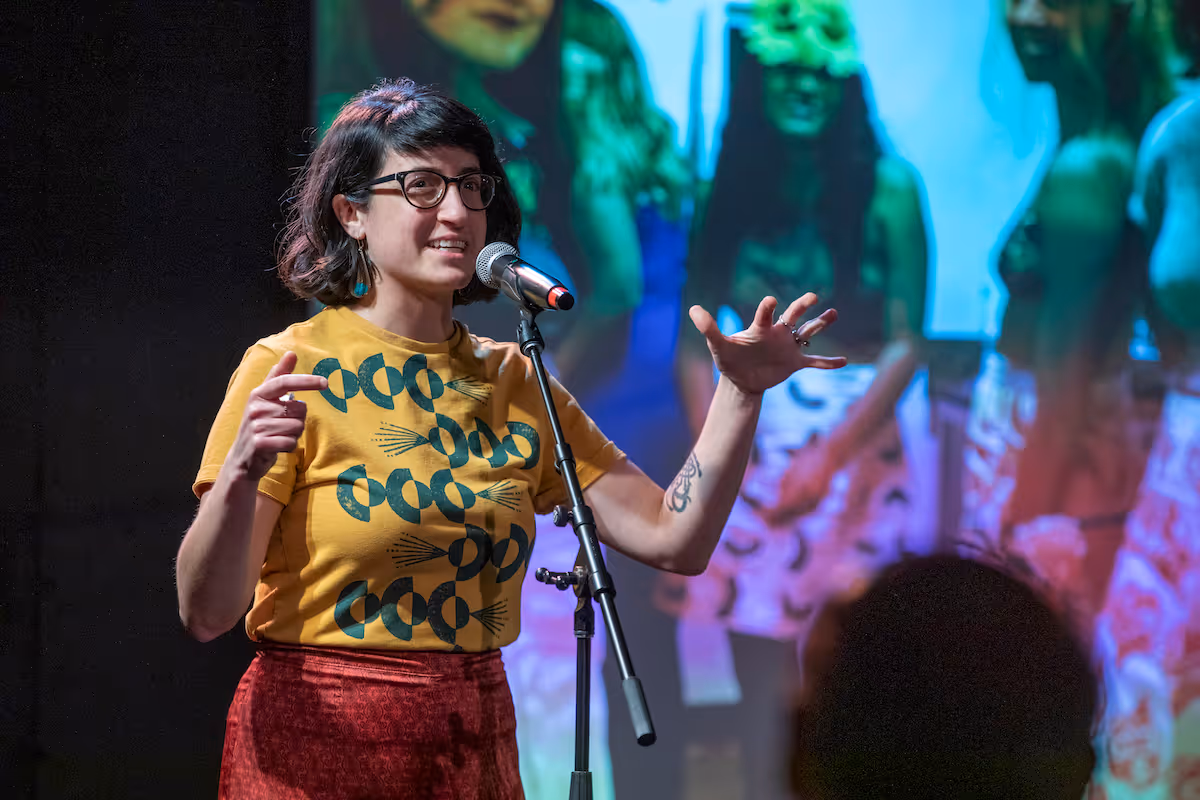 A woman wearing glasses and a mustard yellow shirt with circular designs speaks into a microphone on stage with a colorful projected background.