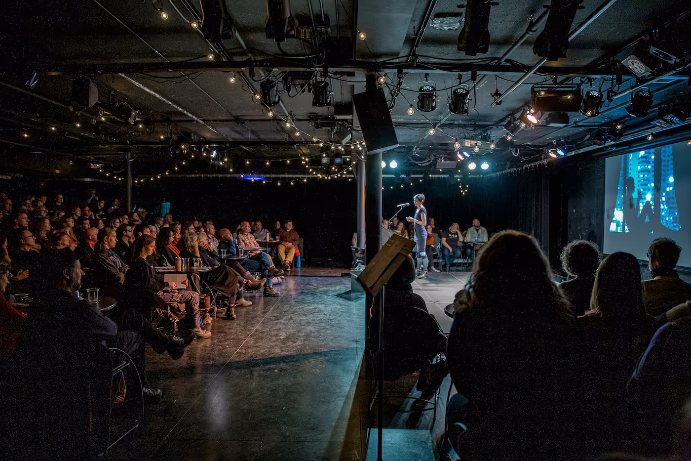 Audience attentively listening to a person speaking on stage in a dimly lit venue with string lights and a projected image on the right.