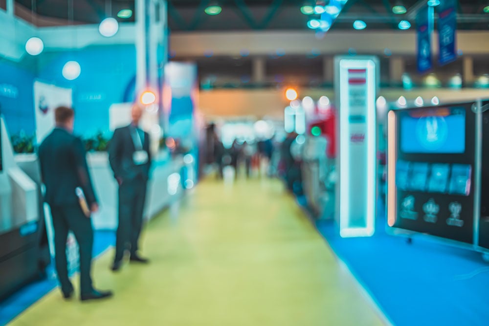 Blurry image of an indoor event or trade show with people standing and walking along a wide aisle with blue and yellow flooring and illuminated displays.