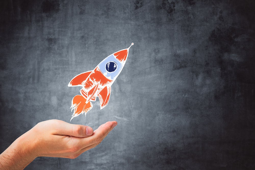 Hand holding a colorful cartoon rocket blasting off against a dark textured background.