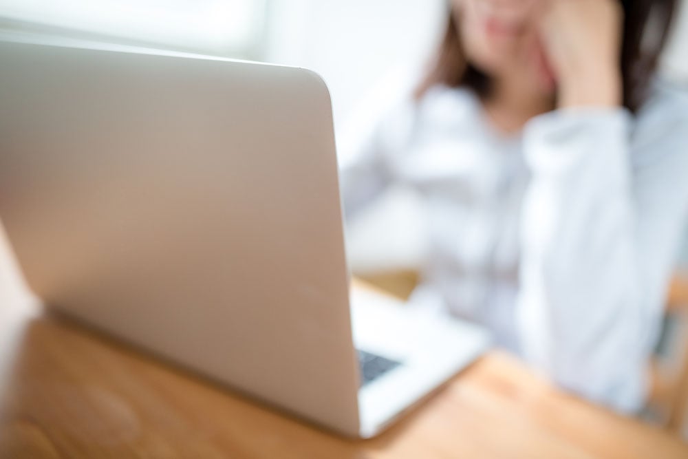 Blurred image of a person sitting at a wooden table using a silver laptop.